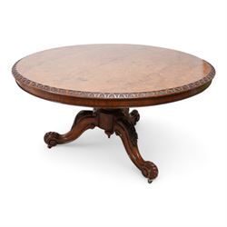 Victorian figured walnut breakfast or centre table, circular tilt-top with carved edge and quarter-matched veneers, turned pedestal with foliate carved baluster on three out splayed supports decorated with floral carvings and scrolled terminals, on brass castors 