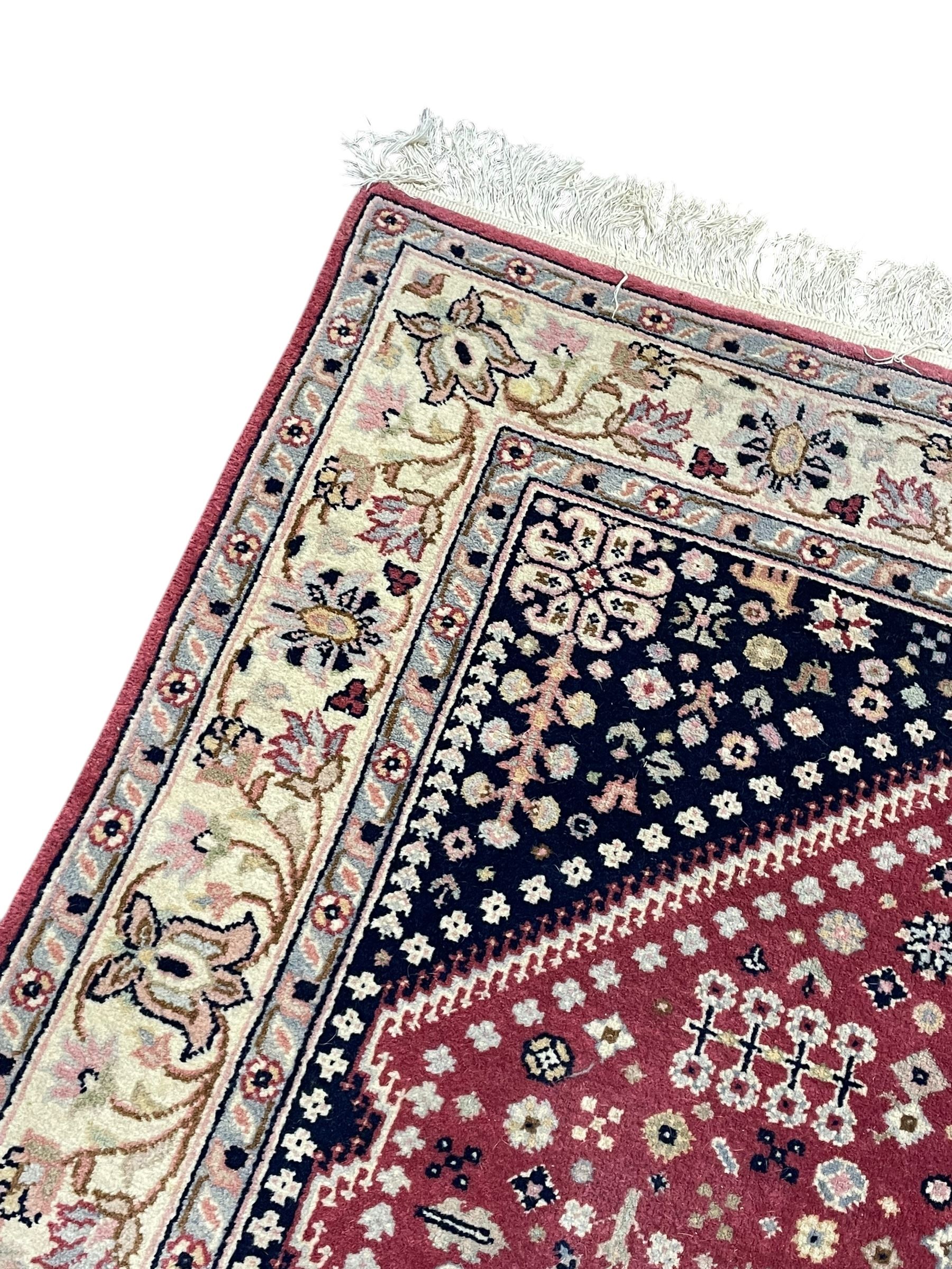 Persian design crimson ground rug, the field decorated with tree of life and floral motifs, enclosed by indigo spandrels with stylised flower head decoration, repeating scrolling border 