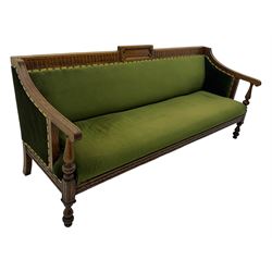 Late Victorian walnut framed sofa, rectangular backrest with carved detailing and central motif, upholstered in olive green fabric with brass studded trim and patterned front rail, scrolled fluted arms supported by turned uprights terminating in bun feet