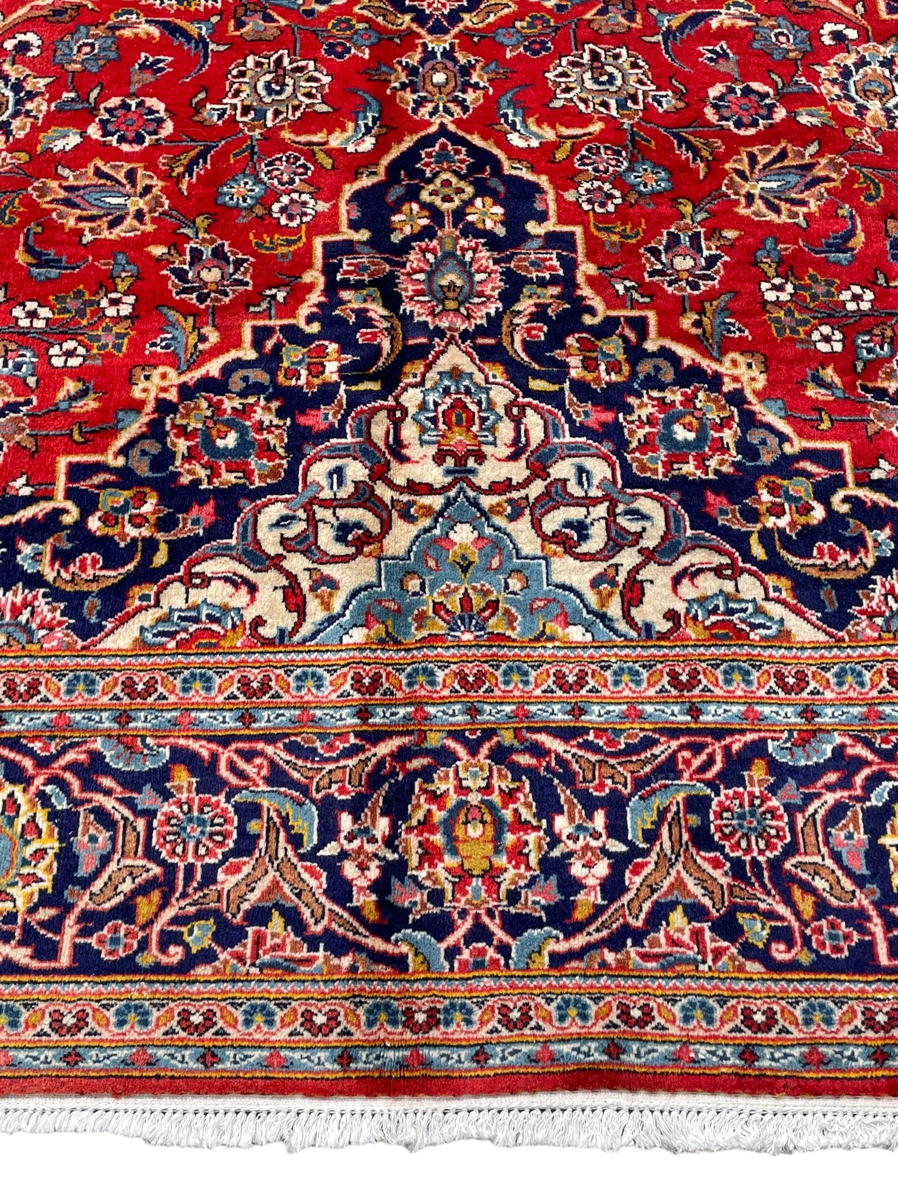 Persian Kashan crimson ground rug, overall arabesque design, the field decorated with swirled leafy branches and palmettes, indigo ground border decorated with repeating pattern, within guard stripes 