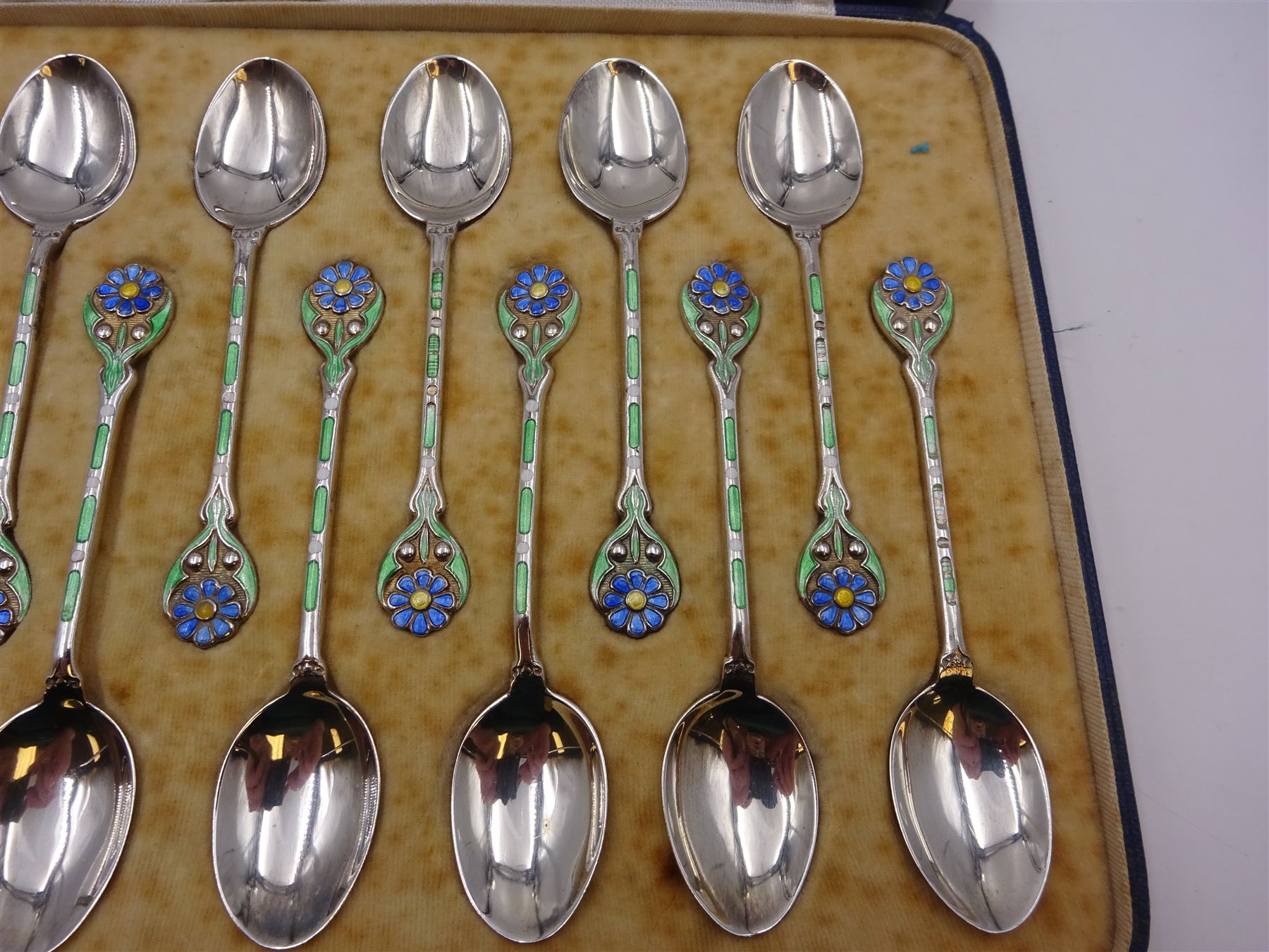 Set of twelve silver coffee spoons, each with enamelled blue flower to terminal and green and white enamel decoration to stem, hallmarked Mappin & Webb Ltd, Birmingham 1934, contained within fitted case