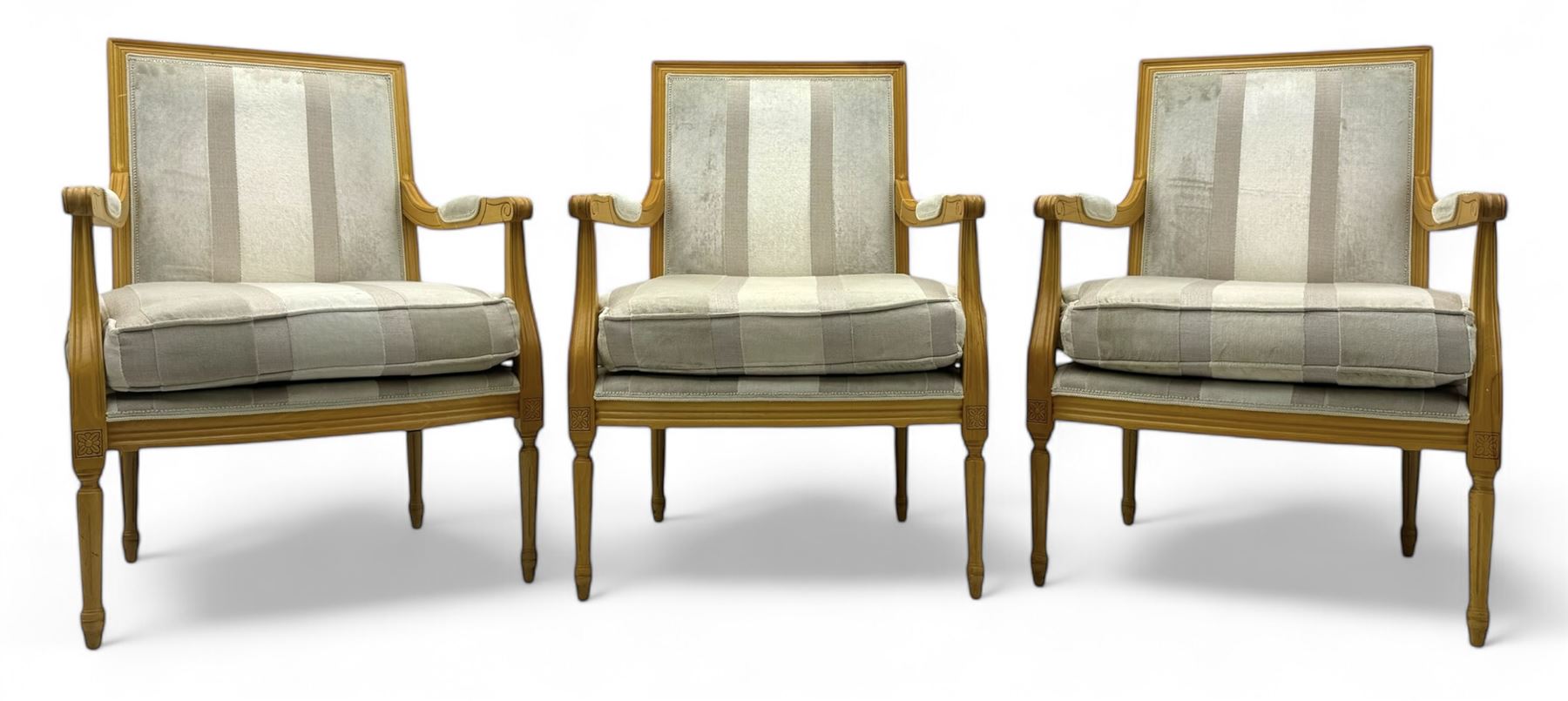 Three mid-20th century beechwood fauteuil armchairs, upholstered backrest and seat in cream stripe fabric, padded armrests with scroll detail, on tapered fluted supports with carved floral rosette blocks