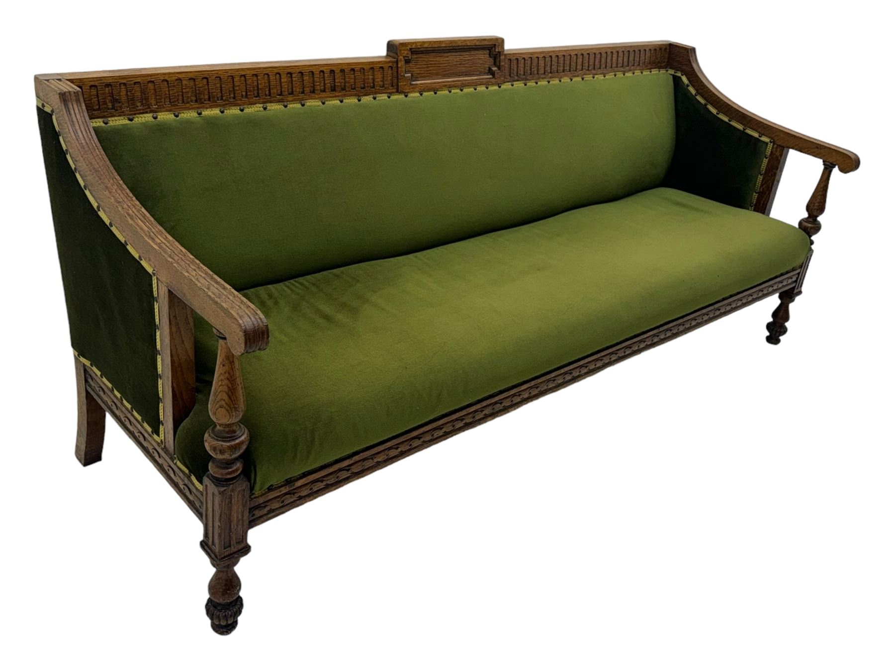 Late Victorian walnut framed sofa, rectangular backrest with carved detailing and central motif, upholstered in olive green fabric with brass studded trim and patterned front rail, scrolled fluted arms supported by turned uprights terminating in bun feet