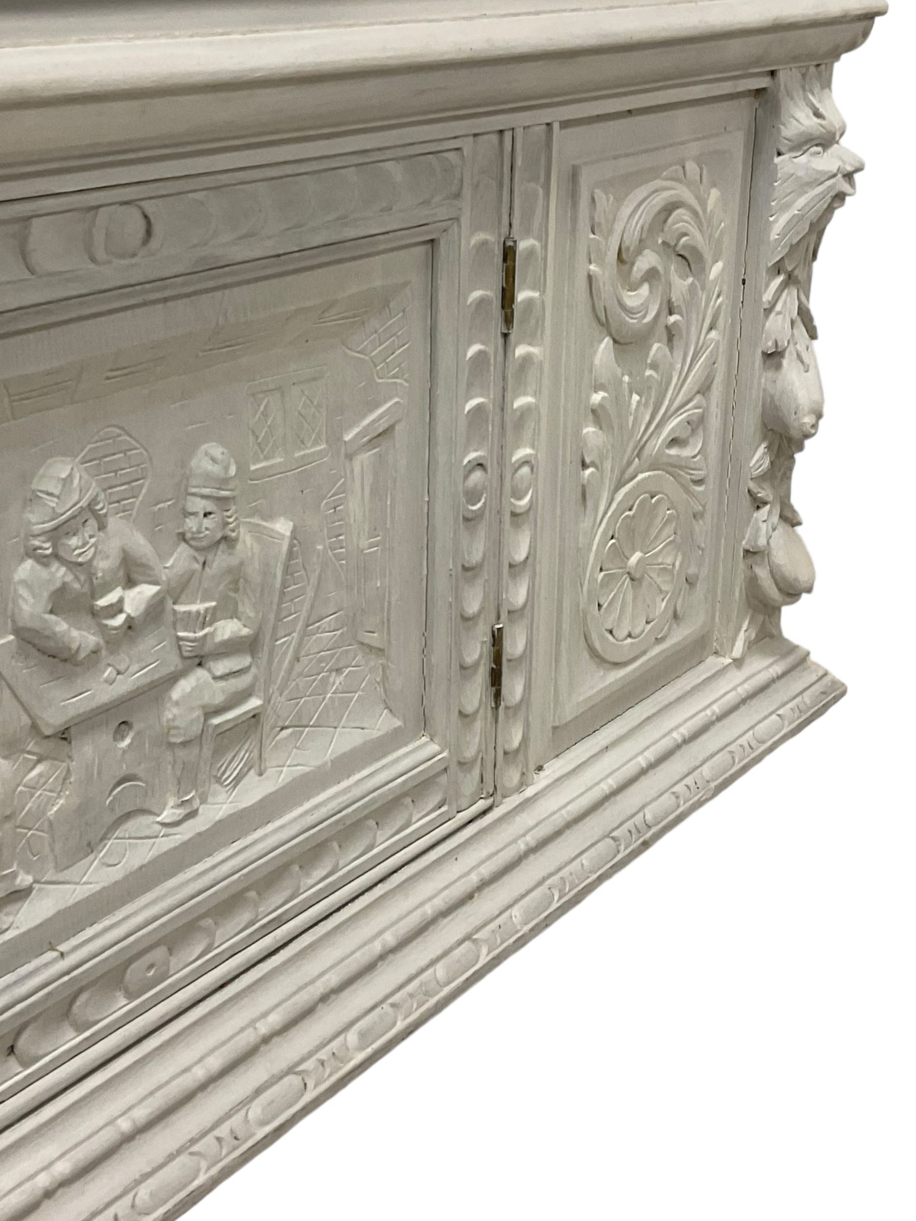 Victorian white painted heavily carved oak buffet, shaped pediment decorated with central urn filled with fruit flanked by winged beasts and scrolling acanthus leaves, moulded rectangular top carved with foliage, two drawers with Greenman mask handles over undertier, the upper tier supported by flute playing musicians, the lower section fitted with cupboard enclosed by panelled door carved with tavern scene, bead carved lower mouldings, on stile supports 