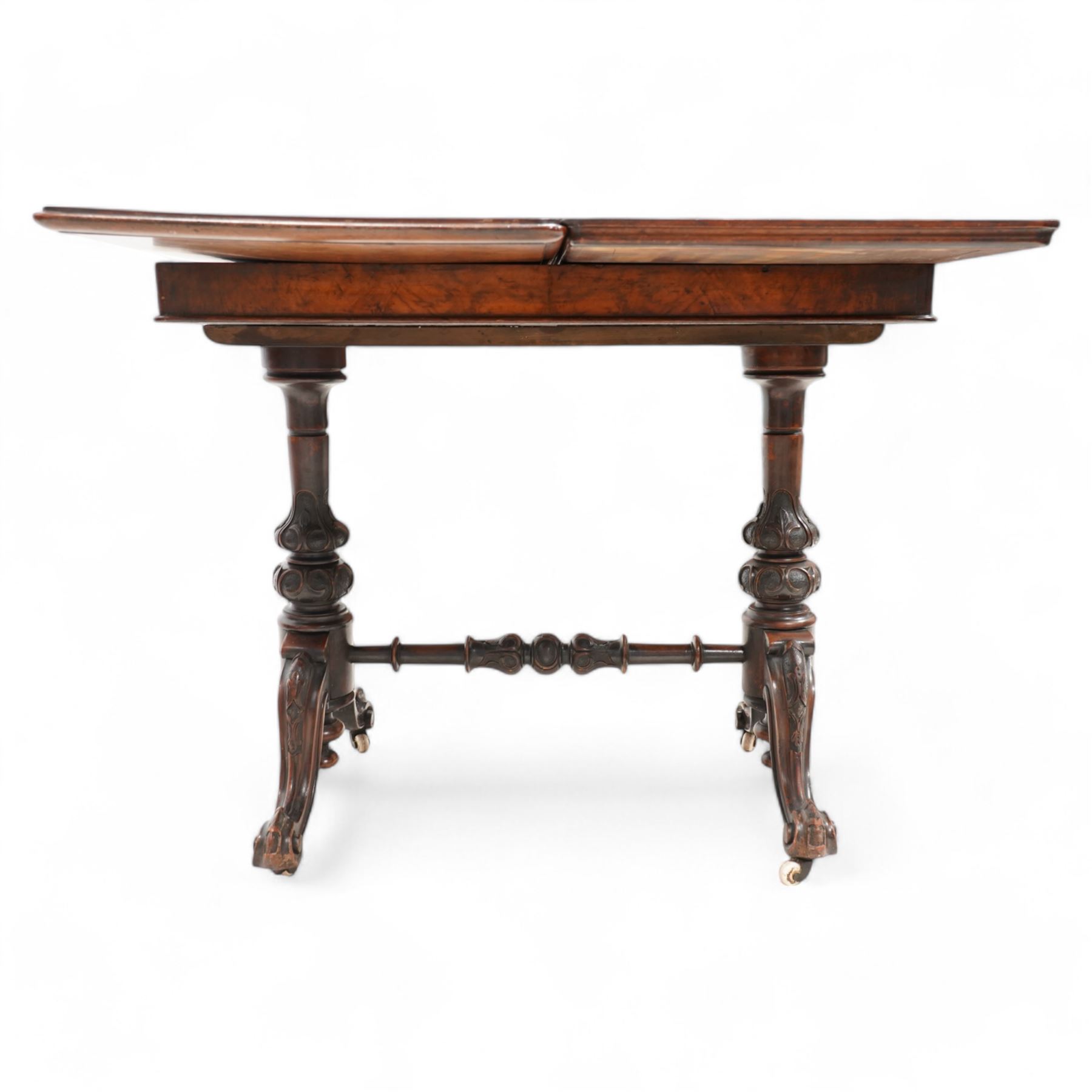 Victorian figured walnut card table, moulded rectangular fold-over swivel top with baize-lined interior, on twin turned pillar supports carved with stylised foliate motifs, united by turned stretcher, on splayed shaped feet with scrolled carved terminals, brass and ceramic castors 