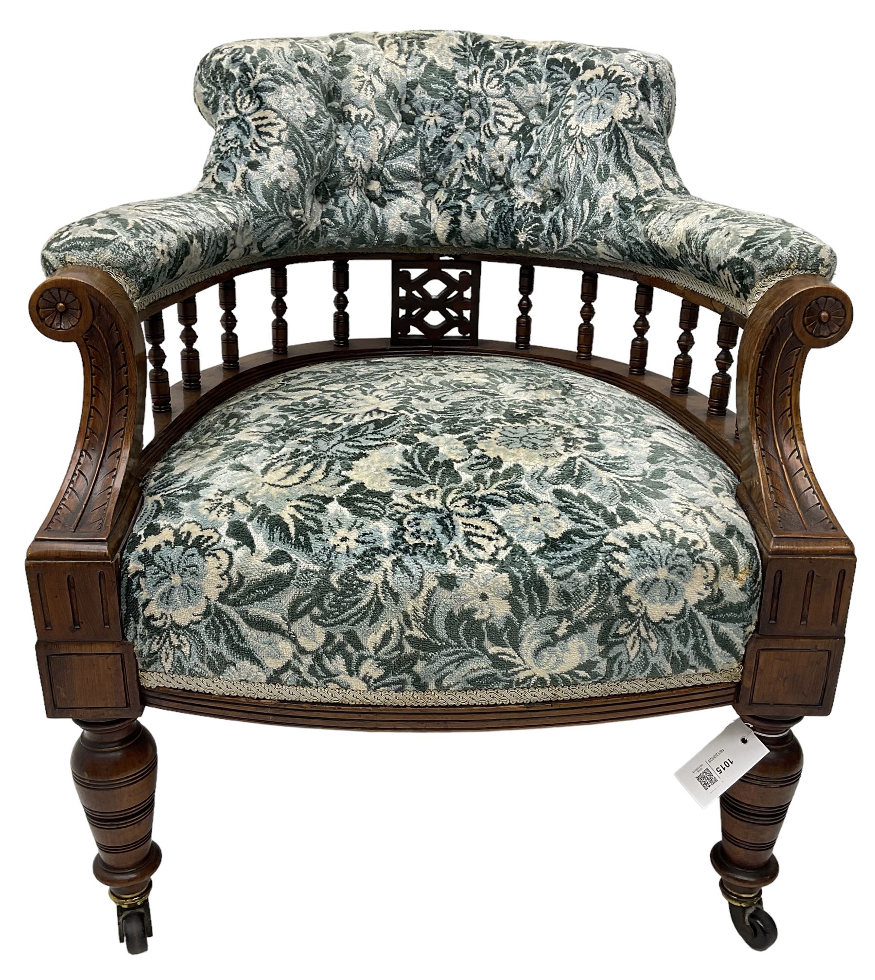 Victorian walnut framed tub-shaped armchair, the shaped rolled back supported by a spindle turned balustrade, upholstered in buttoned floral fabric, scrolled arms supports carved with acanthus leaves and flower heads, on collar turned feet with brass and ceramic castors