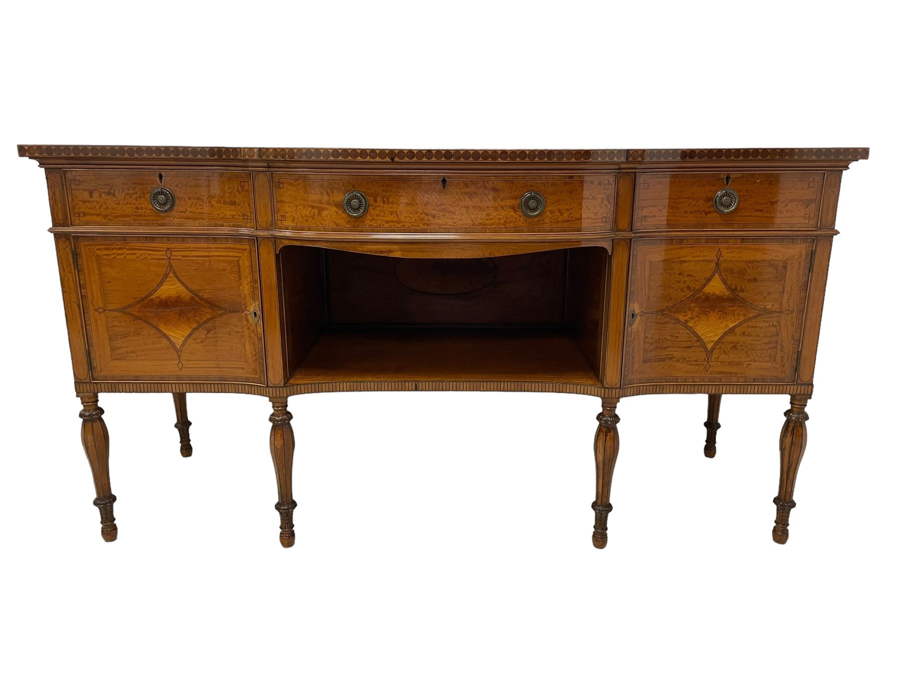 Waring & Gillow - Edwardian Sheraton Revival satinwood sideboard, shaped and stepped break bow-front, the top with central oval book-matched veneered panel and rosewood banding, the front and return edges inlaid with a single row of repeating circles, three frieze drawers each inlaid with rosewood band and ebony stringing, two cupboards flanking open centre, the doors inlaid with lozenges and satinwood cross bandings, on faceted octagonal supports with foliate carved collars, terminating to turned feet, the central drawer with 'Warings' metal plaque and stamped 'Gillows'