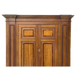 George III and later oak and mahogany banded corner cupboard, moulded cornice over four panelled doors, the door panels inlaid with mahogany bands, projecting square pilasters throughout with applied reeded mouldings, the painted interior fitted with shaped shelves, on moulded plinth base 