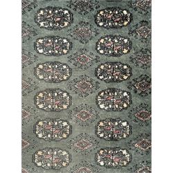 Pakistani Bokhara green ground rug, the field decorated with rows of repeating gul motifs, the main border featuring a series of smaller gul designs and geometric patterns, framed by multiple guard stripes