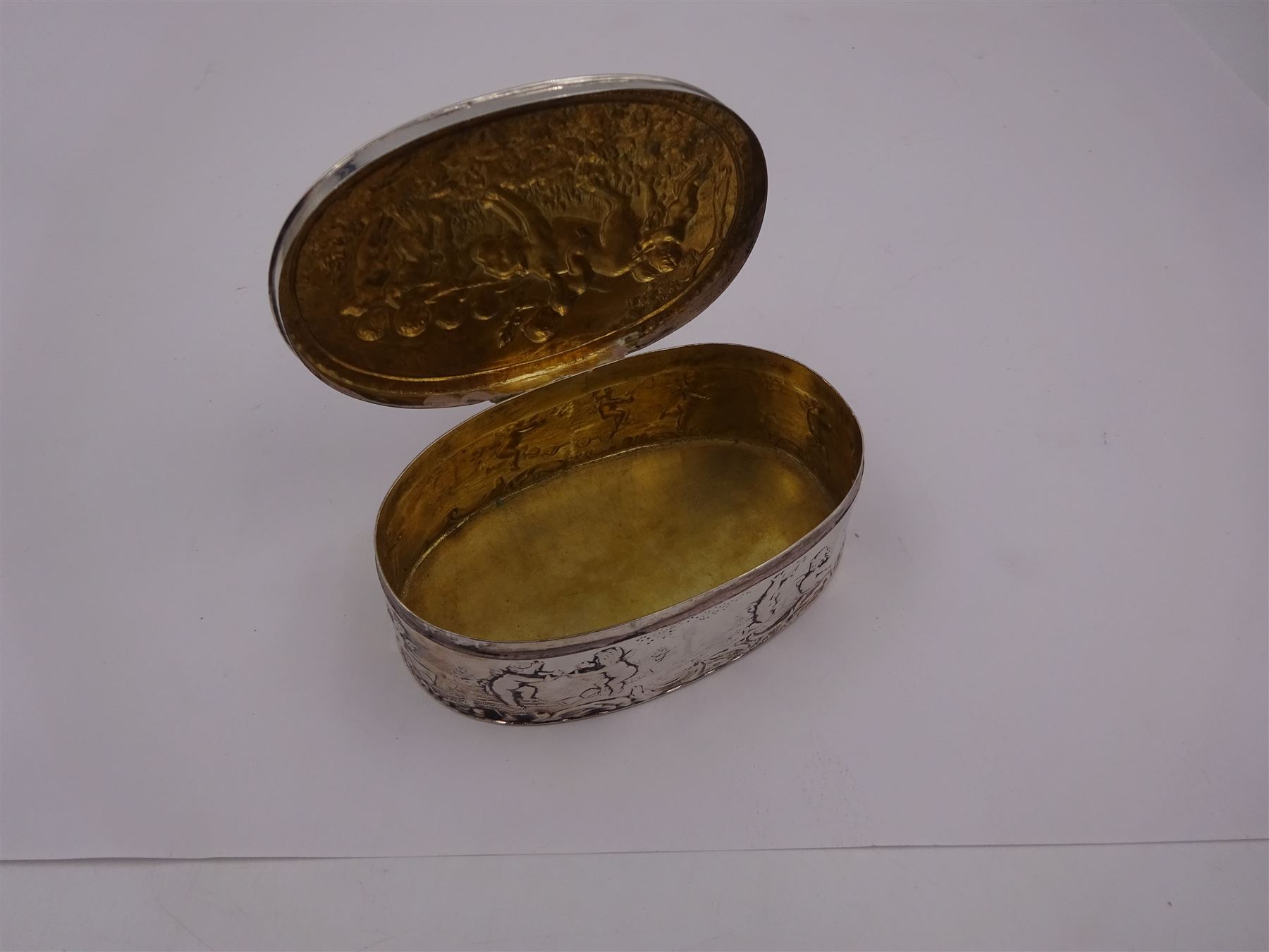 Late 19th century German Hanau silver box, of oval form, embossed throughout with putti in various scenes including riding a dog, playing instruments, pulling a cart and dancing, opening to reveal a gilt interior, with Hanau marks for B Neresheimer & Sohne, W9cm, H3cm