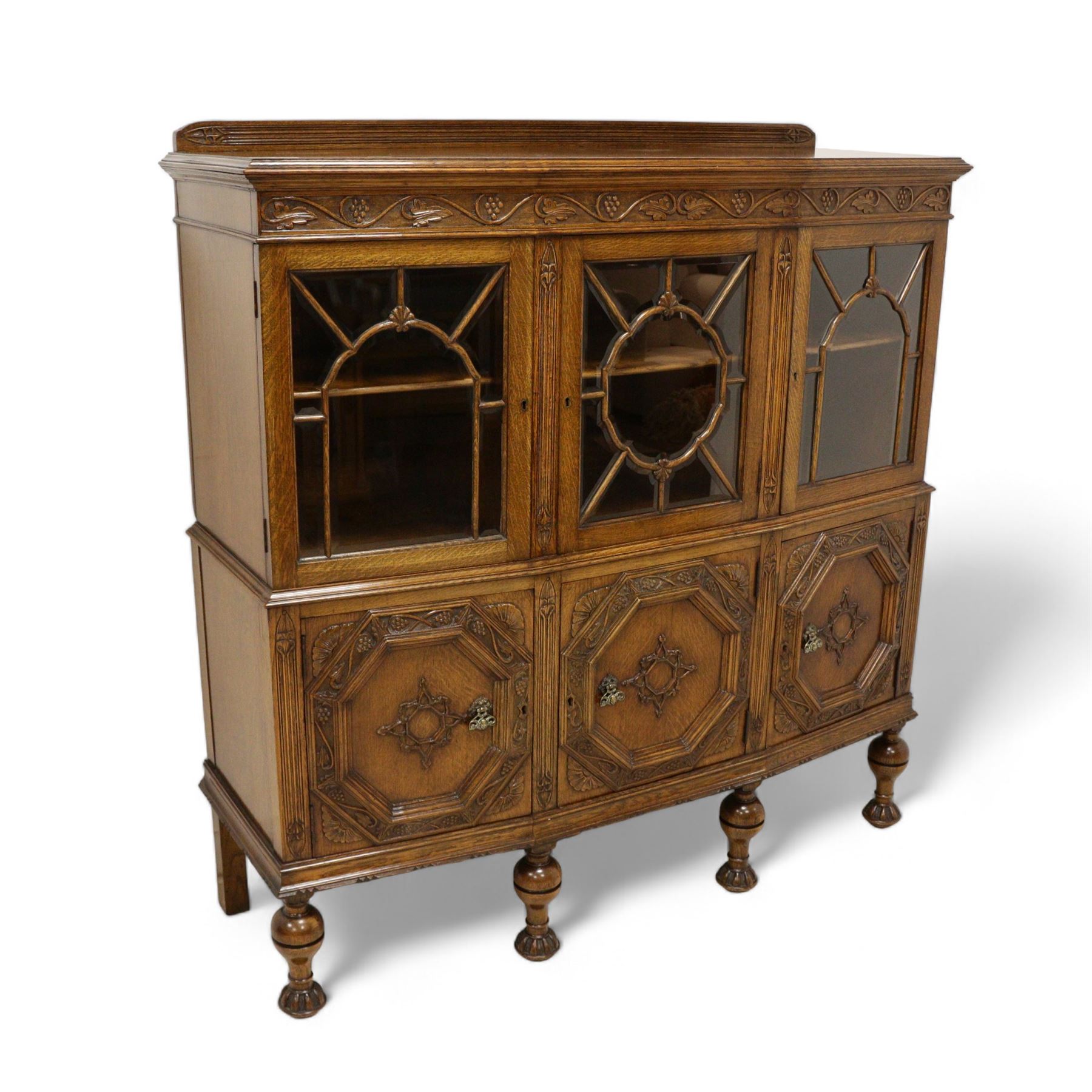 Early 20th century heavily carved oak display cabinet, frieze carved with grape vines over three astragal glazed doors with bevelled glass, over three cupboards applied with octagonal carved panels with further vine carvings, on turned and lobe carved supports