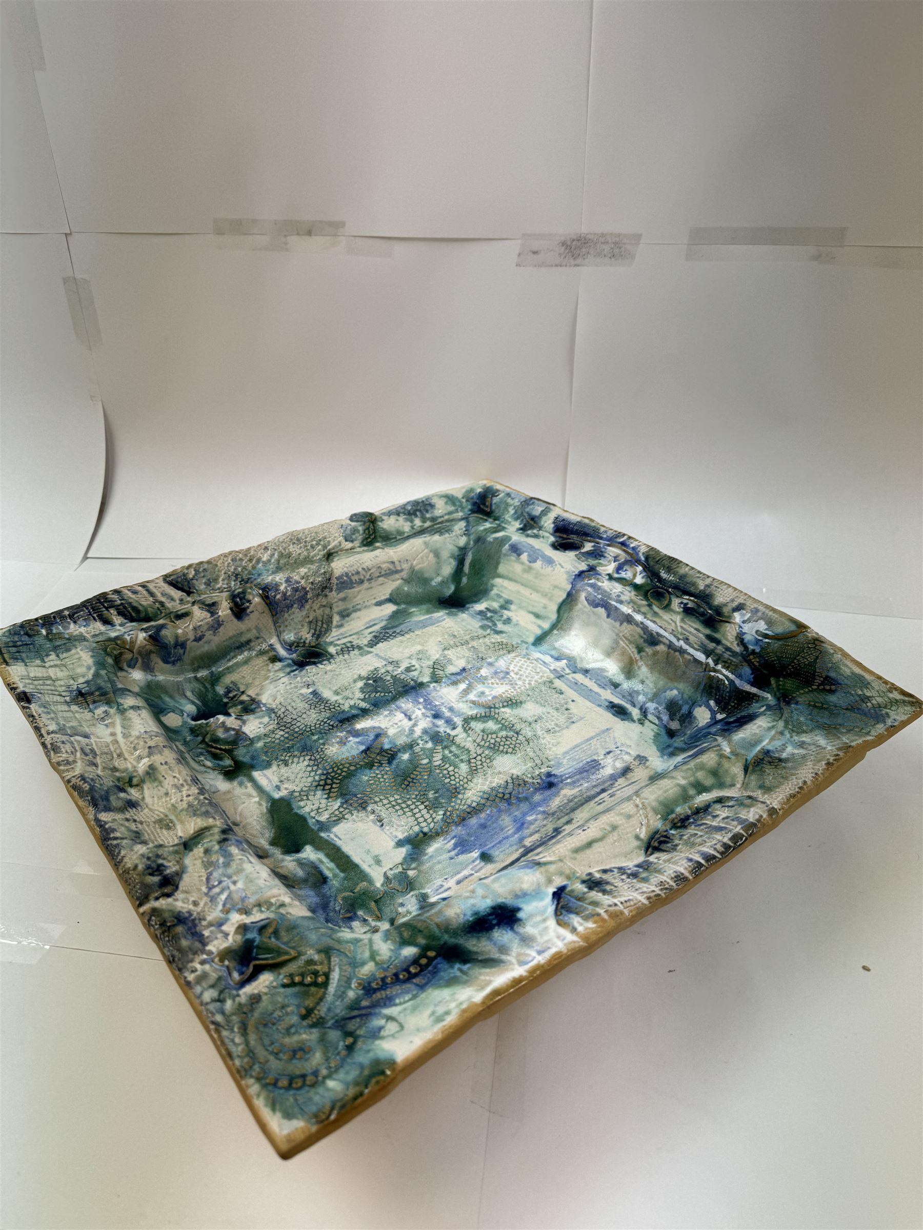 Large studio pottery dish, of square form, with applied and embossed foliate detail on a mottled green/blue ground, W42cm