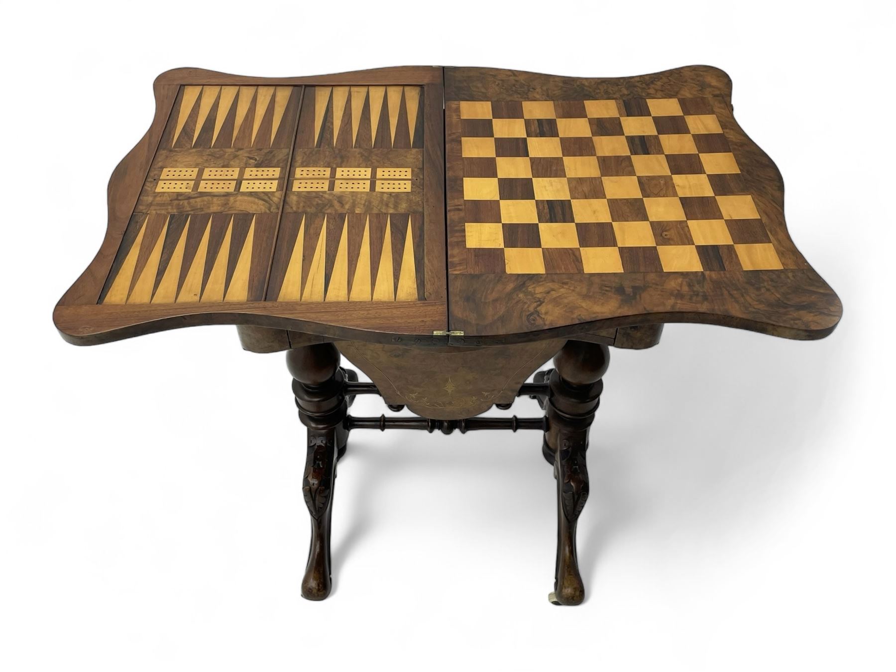 Victorian inlaid figured walnut games table, the serpentine fold-over top inlaid with scrolling foliate decoration, the top opens to reveal cribbage, backgammon and chess boards, single drawer over sliding storage well, on twin pillar supports united by double turned stretchers, on foliage carved out-splayed supports with brass and ceramic castors 