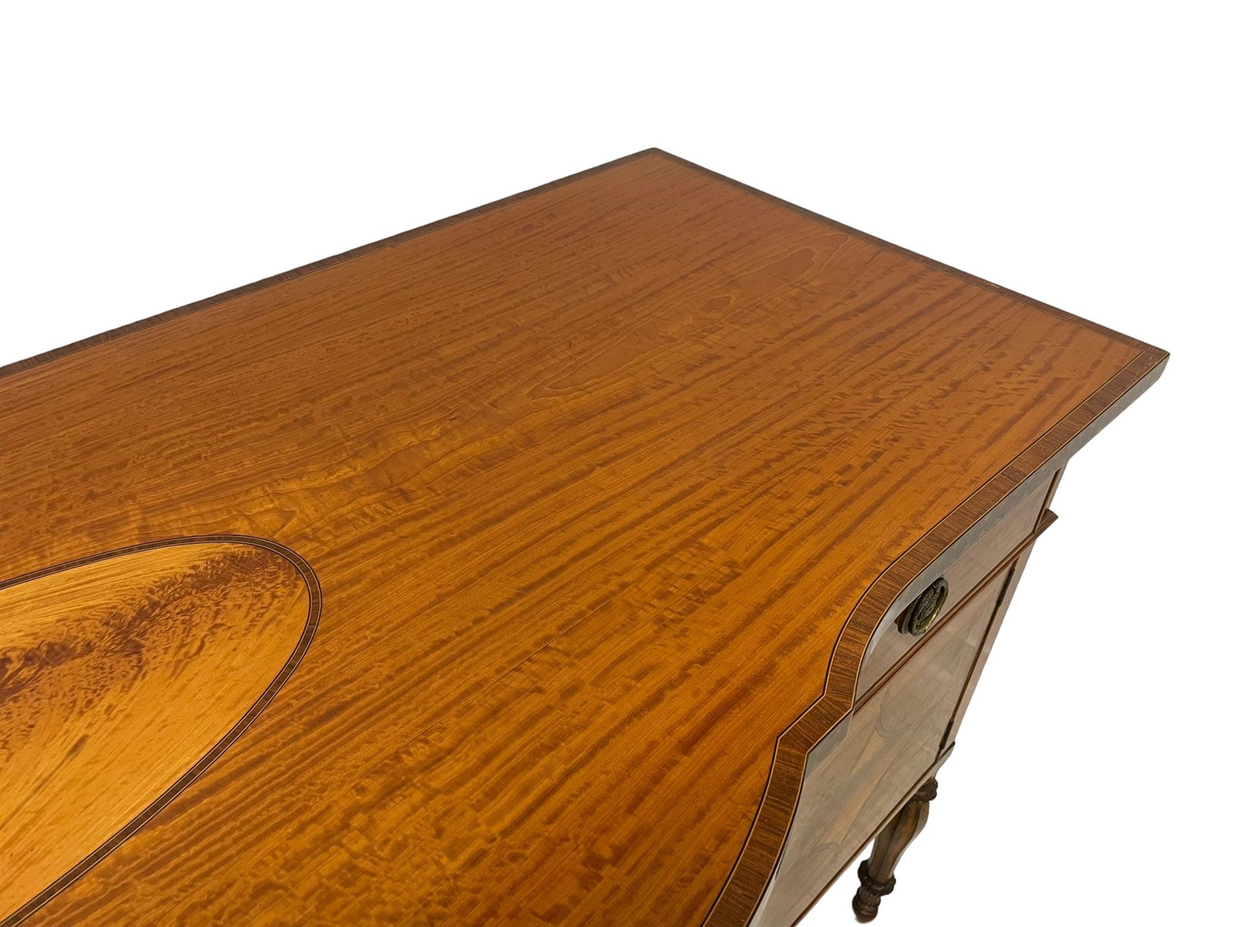 Waring & Gillow - Edwardian Sheraton Revival satinwood sideboard, shaped and stepped break bow-front, the top with central oval book-matched veneered panel and rosewood banding, the front and return edges inlaid with a single row of repeating circles, three frieze drawers each inlaid with rosewood band and ebony stringing, two cupboards flanking open centre, the doors inlaid with lozenges and satinwood cross bandings, on faceted octagonal supports with foliate carved collars, terminating to turned feet, the central drawer with 'Warings' metal plaque and stamped 'Gillows'