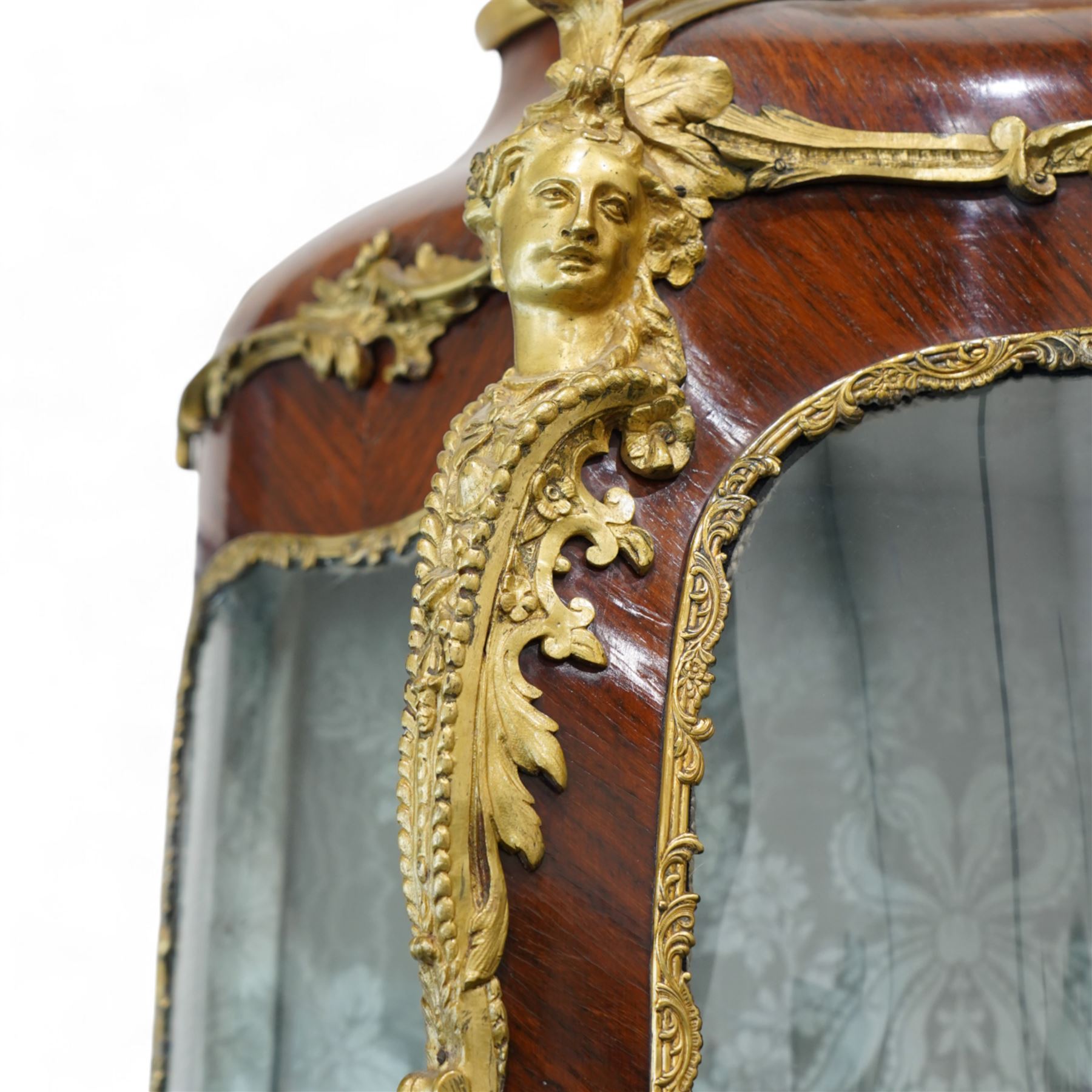 Large early 20th century French gilt metal mounted vitrine, serpentine and bombe form, C-scroll pediment decorated with extending flower heads, shaped front enclosed by single glazed door and glazed with shaped glass panes with foliate cast edging, floral pattern silk lined interior fitted with two glass shelves and one lined shelf, hand painted with Vernis Martin design panels depicting garden courting scene and putti, on cabriole supports mounted by shell and C-scroll mounts 