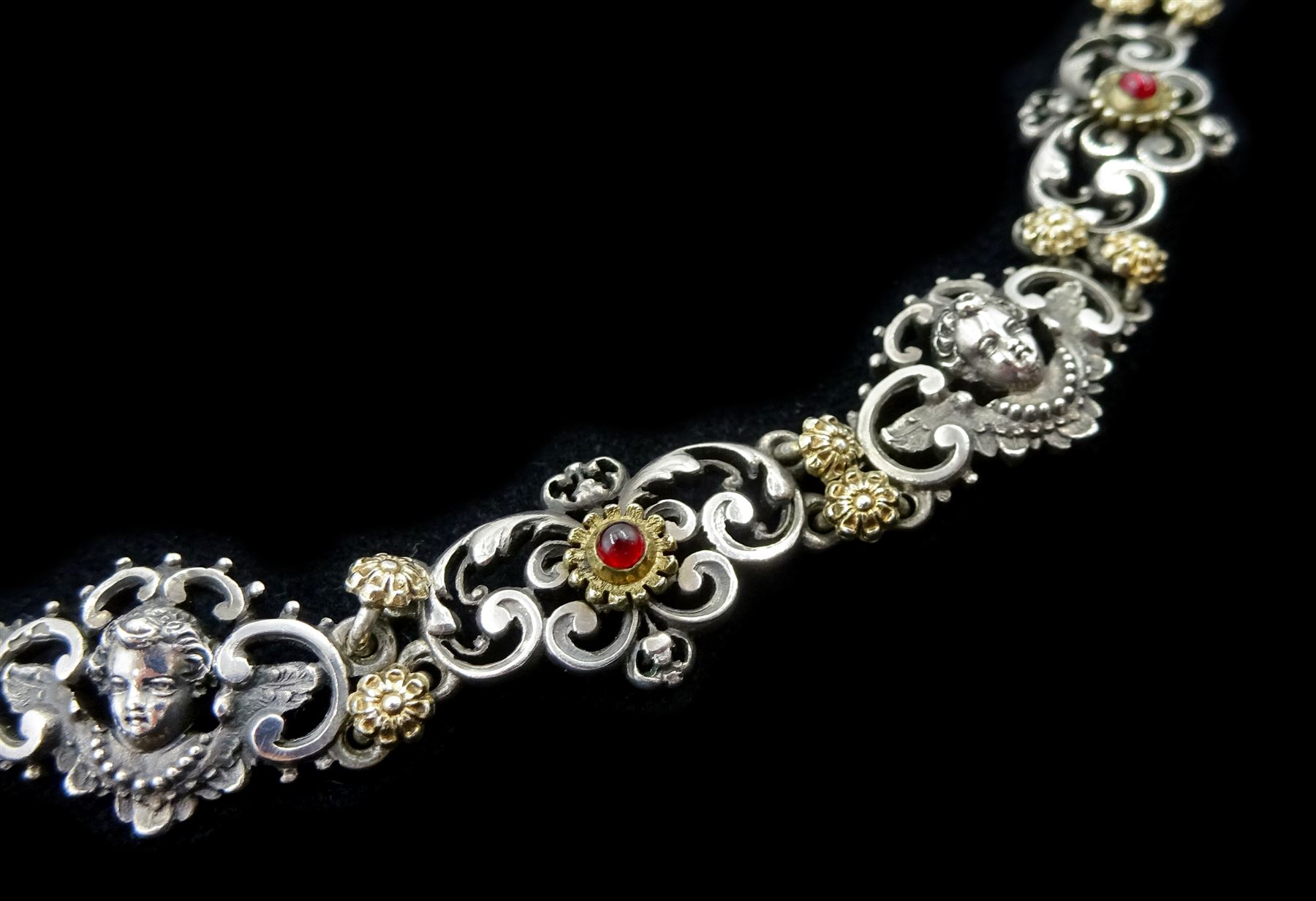 Early 20th century continental silver scroll link necklace, alternate panels of winged cherub and silver-gilt cabochon red paste stones, separated by silver-gilt flower head links, indistinct hallmarks, possibly Austrian or German