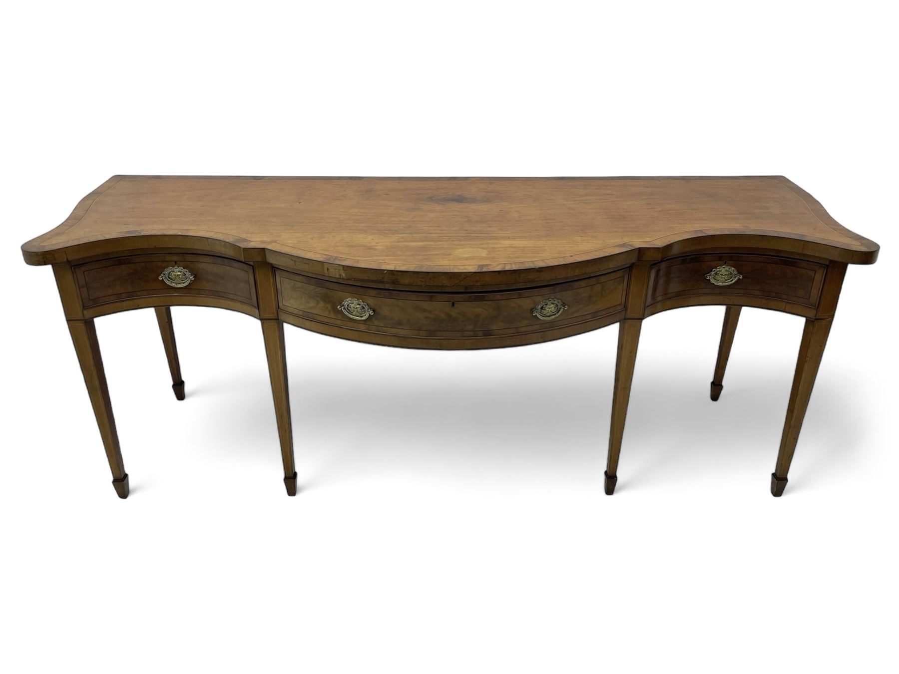 Large George III mahogany serpentine serving table, shaped top with crossing banding and stringing, the frieze fitted with three cock-beaded drawers, oval pressed brass handle plates decorated with urns, on square tapering supports with spade feet 