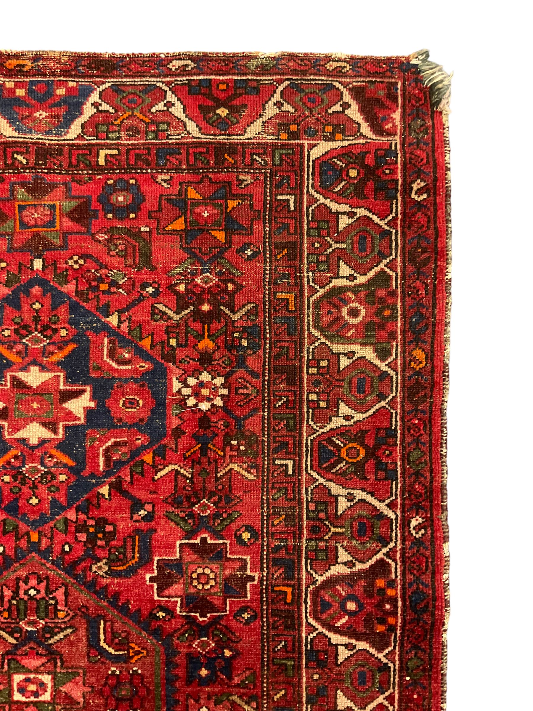 Persian Hamadan rug, red ground with overall geometric design, the field with three extended lozenges, decorated with geometric and stylised flower head motifs, the border and guards decorated with repeating geometric motifs