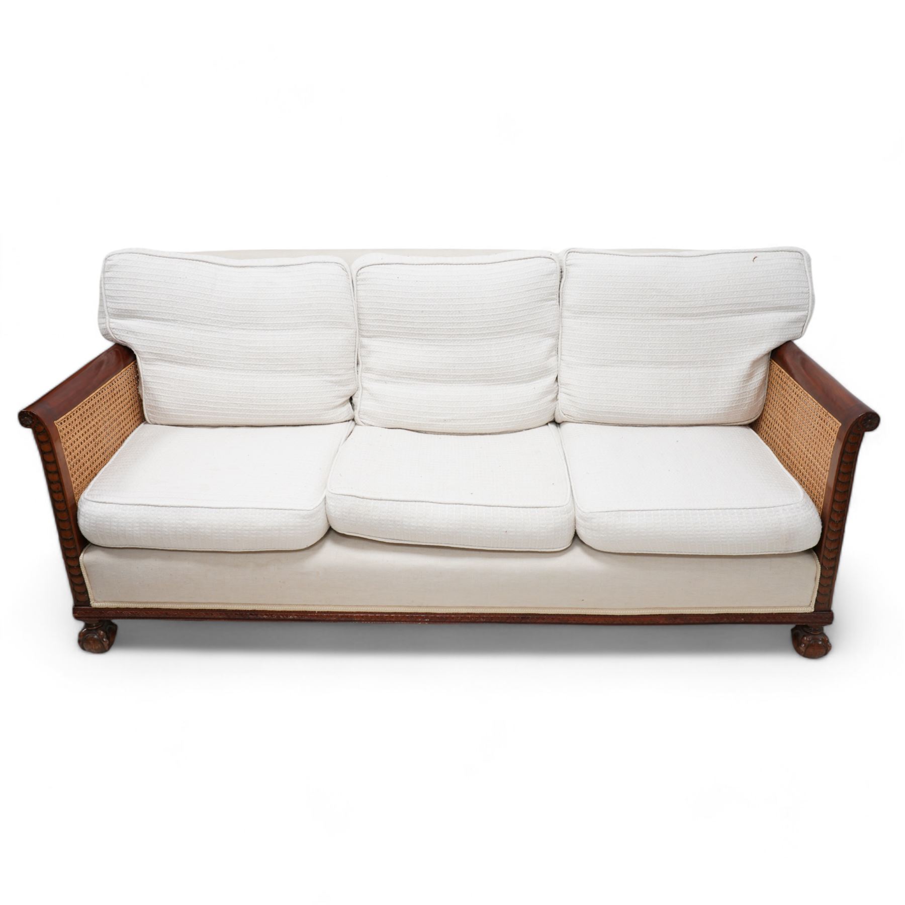 Early 20th century mahogany framed bergère three seat sofa, upholstered back and seat flanked by rolled double-caned arms with carved scale decoration, floral and ribbon-twist carved lower edge, over ball-and-claw feet