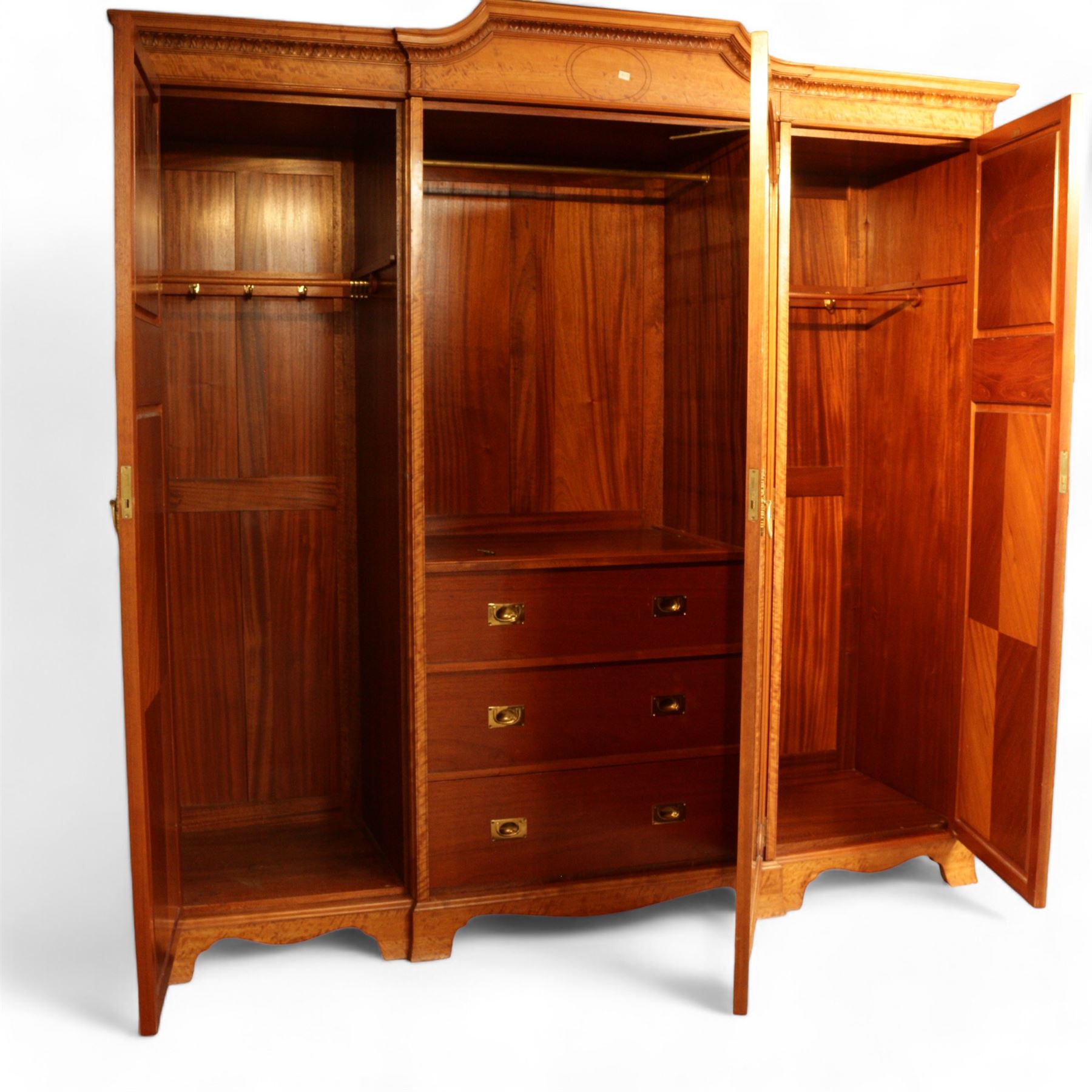 Waring & Gillow Ltd. - Edwardian inlaid satinwood triple wardrobe, stepped moulded cornice over foliate carved edge, single bevelled mirror glazed door enclosing hanging rail and three drawers, flanking panelled doors enclosing hanging rails and hooks, the door frames and panels inlaid with geometric stringing in ebony and satinwood lozenges, reeded and foliate cast loop handles and foliage cast handle plate escutcheon, quarter-matched veneers, on shaped bracket base, the locks stamped 'Gillows Lancaster' and the right hand door with metal marker's plaque 