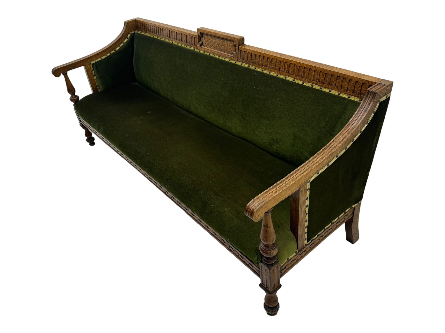 Late Victorian walnut framed sofa, rectangular backrest with carved detailing and central motif, upholstered in olive green fabric with brass studded trim and patterned front rail, scrolled fluted arms supported by turned uprights terminating in bun feet