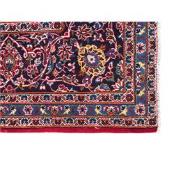 Large Kashan crimson ground carpet, central indigo medallion with stylised floral motifs, flanked by matching spandrels, bordered by multiple bands featuring repeating foliate and floral designs throughout