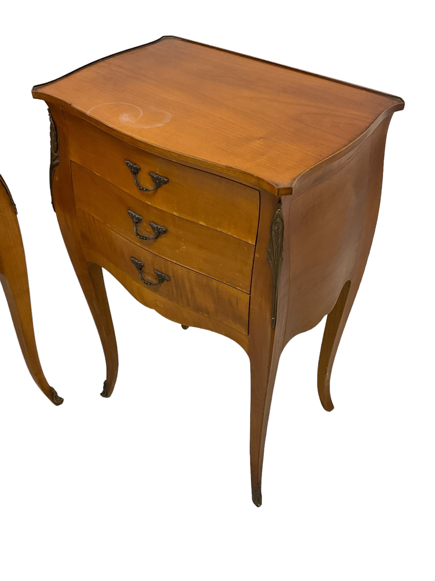 Pair of late 20th century French design cherry wood bedside lamp tables, shaped bombe form, fitted with three drawers, on tapering cabriole supports, decorated with ornate cast gilt metal mounts 