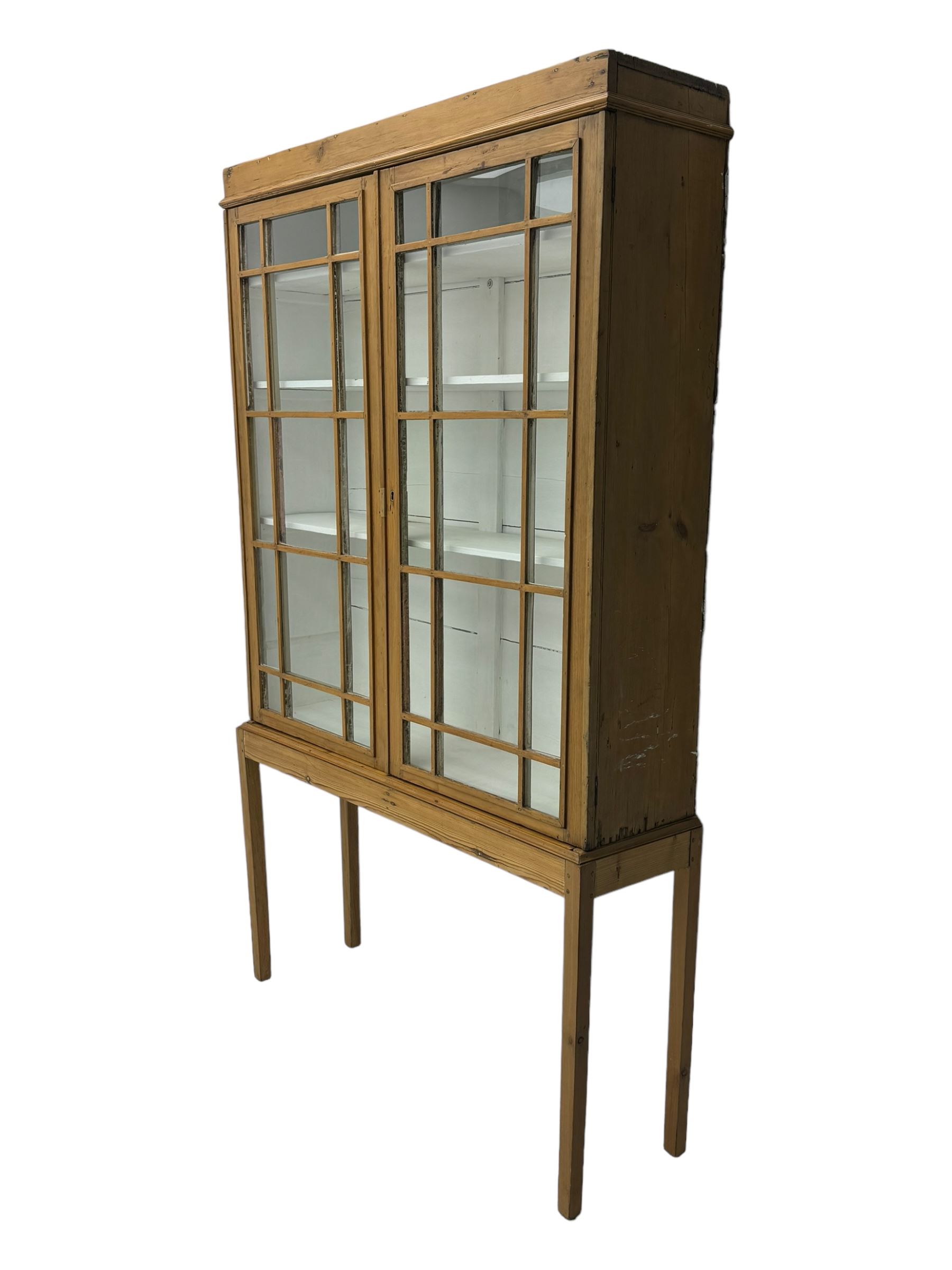 Georgian stripped pine bookcase display cabinet, plain moulded cornice over two glass-panel doors with multiple panes, enclosing white painted interior with three shelves, raised on square supports