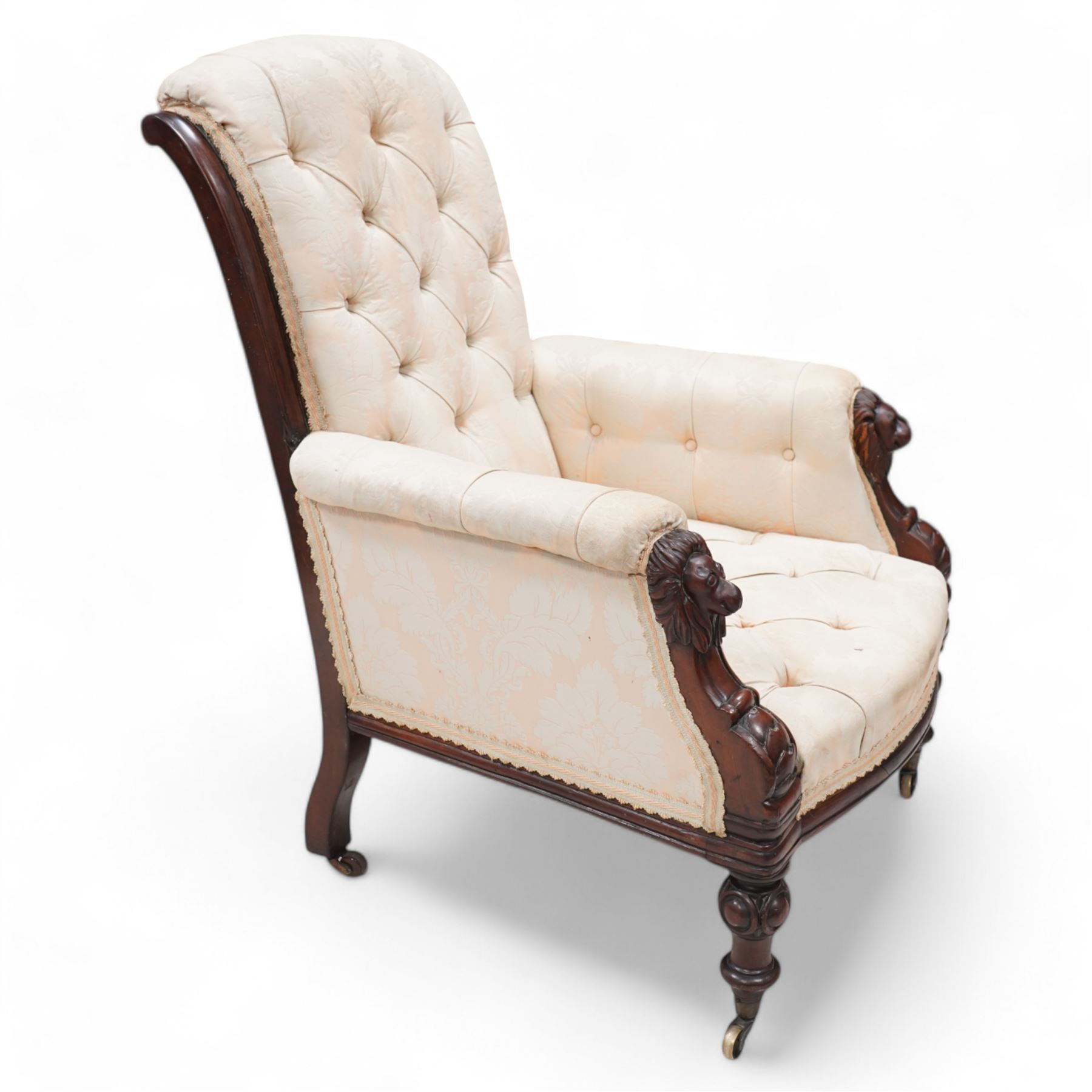 Victorian mahogany framed armchair, moulded and rolled uprights, upholstered in buttoned cream foliate pattern fabric, carved lion mask arm terminals, on turned and globular carved front feet, brass castors 