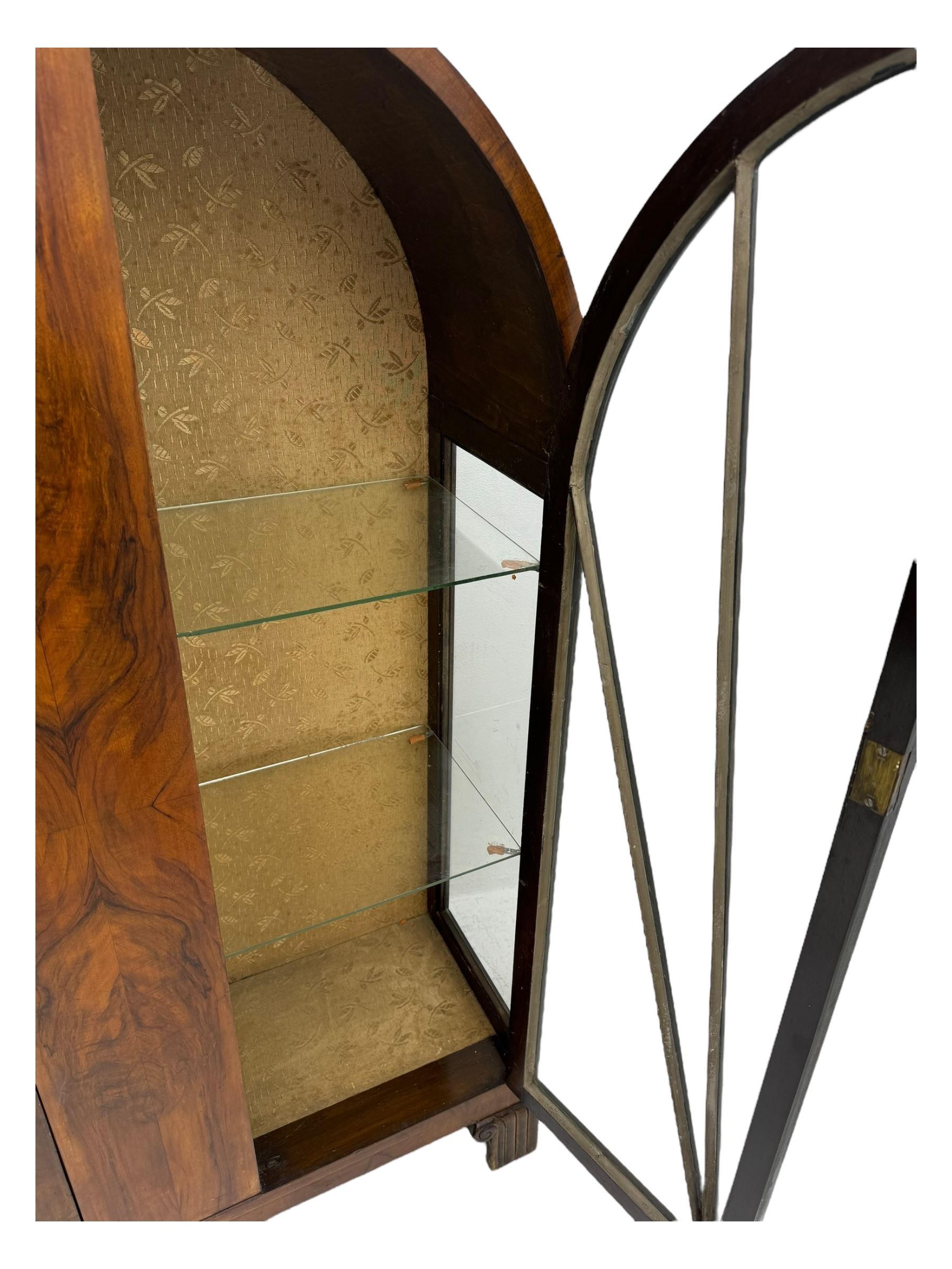 Early 20th century Art Deco walnut display cabinet, with arched top and carved floral crest, central band flanked by two glazed panel doors with decorative mullions, enclosing two glass shelves, on scroll carved bracket feet