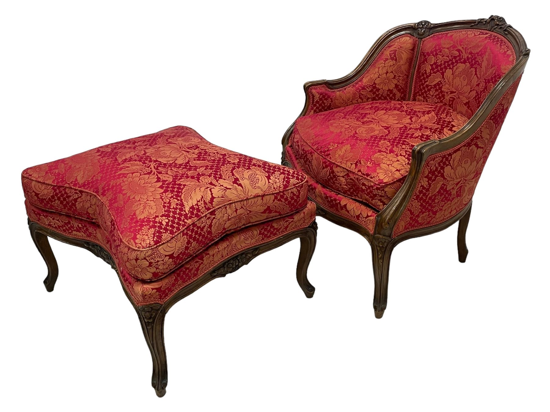 Late 20th century French design stained beech framed armchair and matching stool - the armchair with shaped and moulded frame carved with flower heads and foliage, upholstered in silk red and gold ground fabric with floral pattern, on scroll carved cabriole feet decorated with flower head carvings (W73cm, H82cm, D75cm); the footstool of concaved rectangular form with moulded frame carved with flower heads, upholstered loose cushion, on cabriole feet (70cm x 64cm, H48cm)