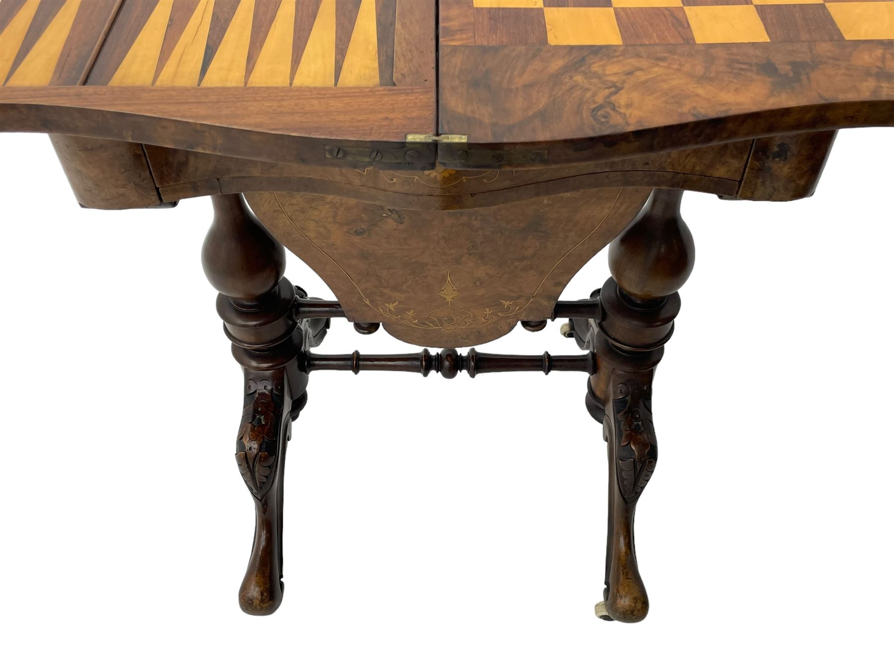 Victorian inlaid figured walnut games table, the serpentine fold-over top inlaid with scrolling foliate decoration, the top opens to reveal cribbage, backgammon and chess boards, single drawer over sliding storage well, on twin pillar supports united by double turned stretchers, on foliage carved out-splayed supports with brass and ceramic castors 