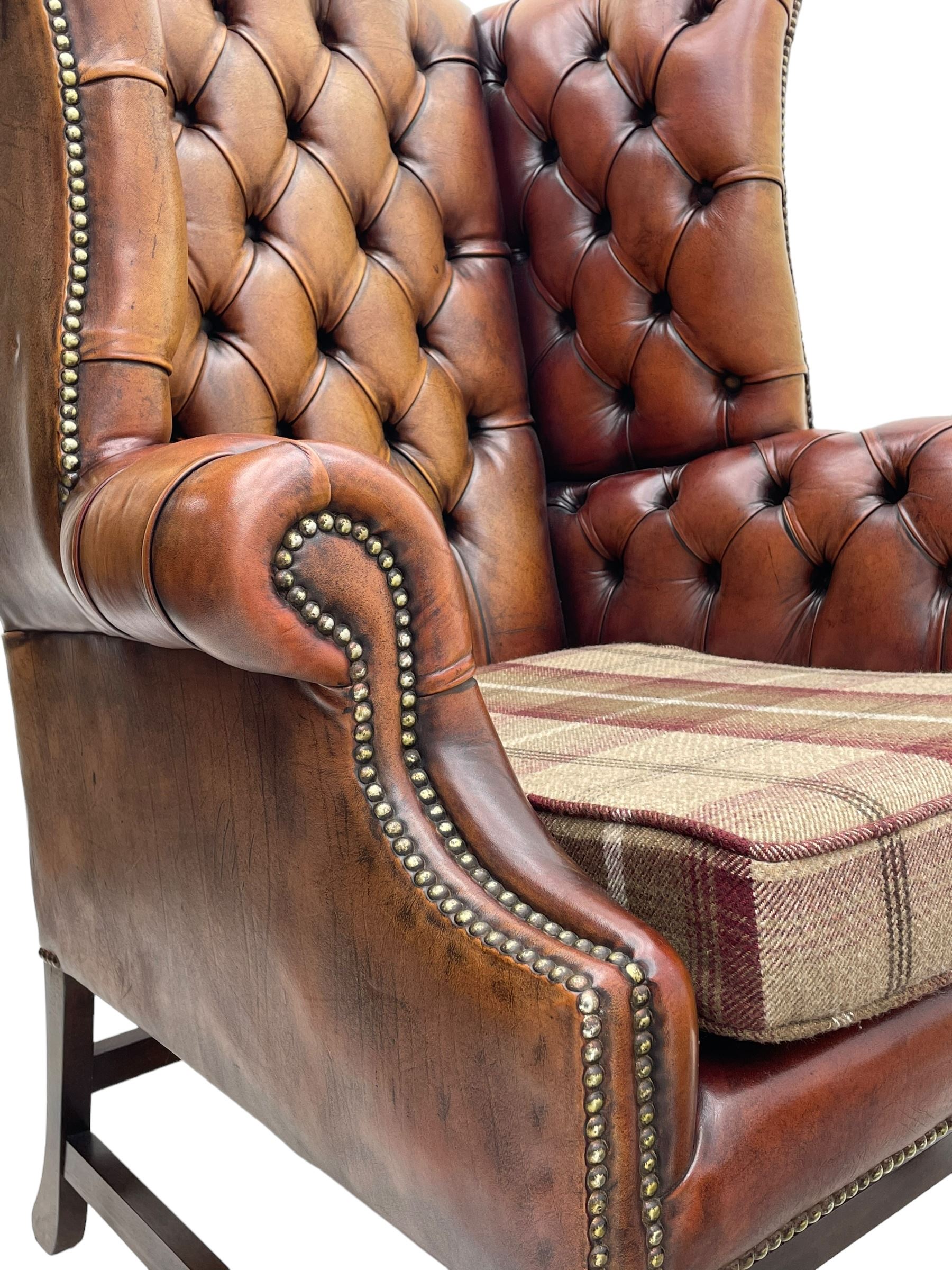 Georgian design hardwood-framed wingback armchair, shaped cresting rail and deep wingback over rolled arms, upholstered in brown buttoned leather with studwork bands, the seat cushion upholstered in checkered fabric, on square supports united by stretchers (W86cm, H118cm, D86cm); together with matching rectangular footstool (63cm x 43cm, H37cm) 