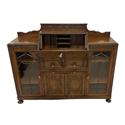 Early 20th century heavily carved oak bookcase bureau, raised back with grape-vine carved frieze, over fall-front enclosing fitted interior, above two short drawers and curved frieze drawer, flanked by astragal glazed and panelled cupboards, on turned feet