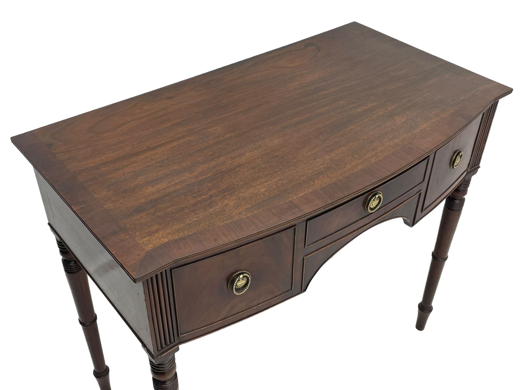 George III mahogany kneehole dressing or writing table, stepped bow-front form, reed moulded top with mahogany crossband, fitted with three cock-beaded drawers with circular pressed brass handle plates decorated with urn and beaded ring handle, on ring turned supports 