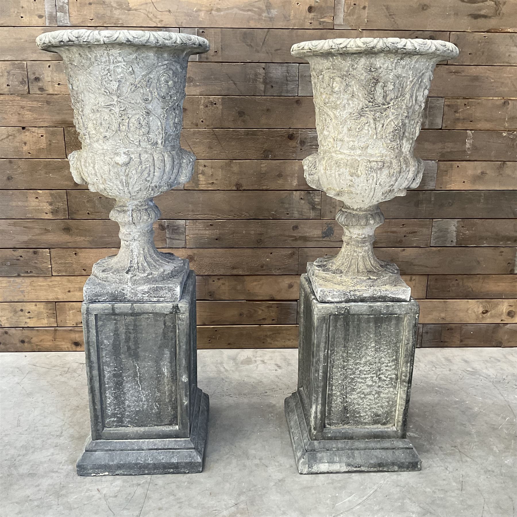 Pair of Georgian design cast stone garden urns, egg and dart rim with raised floral motifs, pedestal base terminating on square plinth, raised on square block plinth