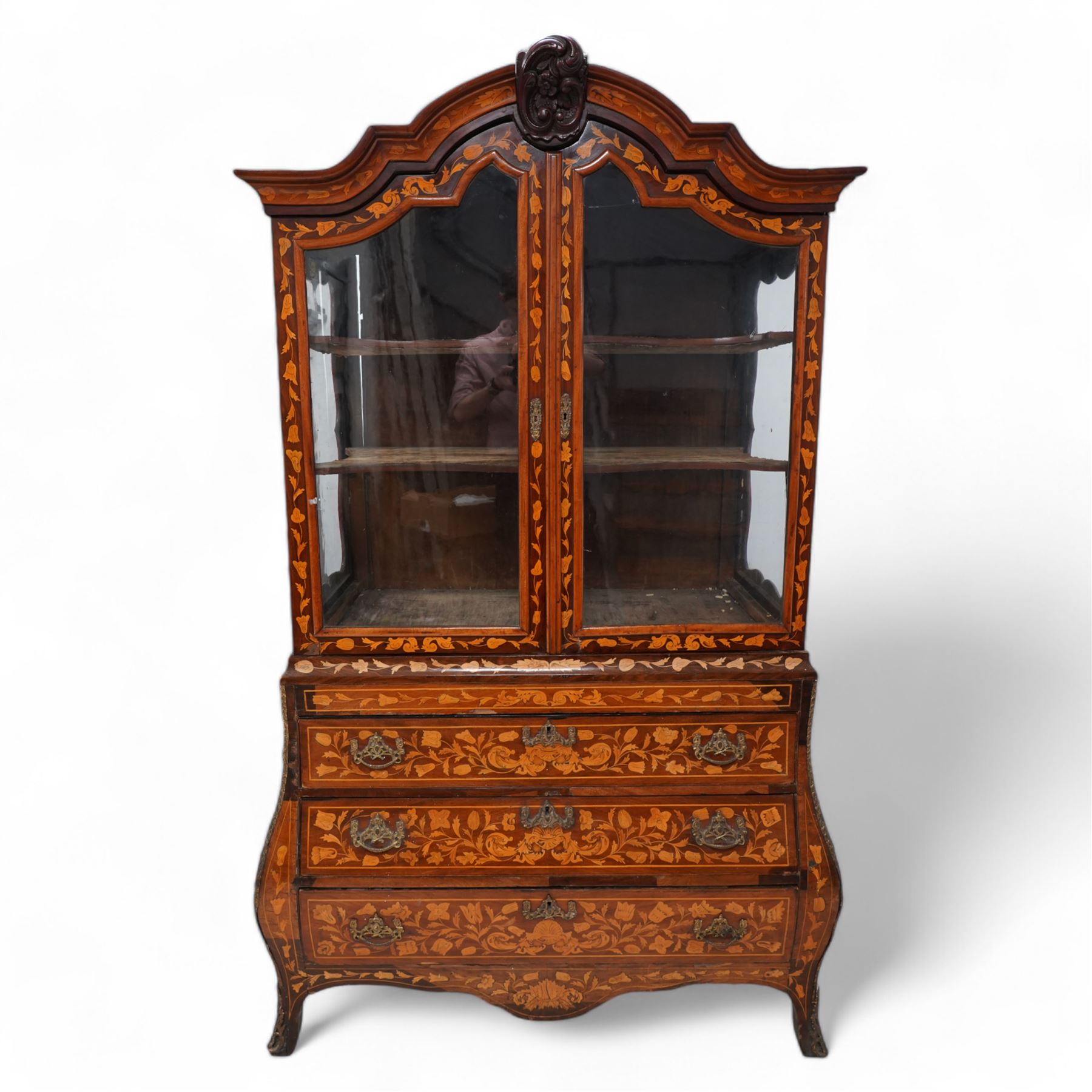 18th century Dutch marquetry walnut and gilt metal mounted vitrine cabinet on chest, shaped pediment carved with scrolling foliage cartouche, inlaid with trailing leafy branches and flower heads, two internal shelves enclosed by two glazed doors, the lower section with bombe outline fitted with three drawers, with ornate cast gilt metal edging and terminal caps