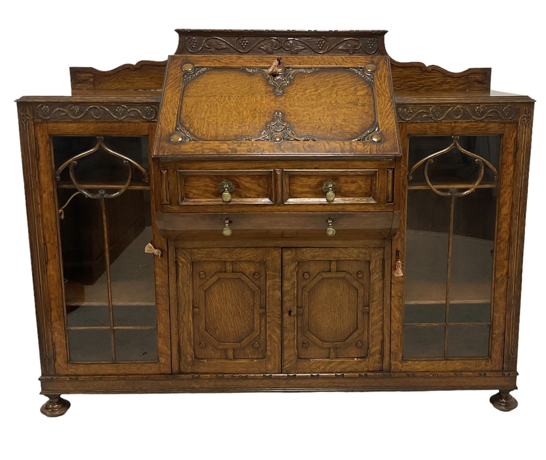 Early 20th century heavily carved oak bookcase bureau, raised back with grape-vine carved frieze, over fall-front enclosing fitted interior, above two short drawers and curved frieze drawer, flanked by astragal glazed and panelled cupboards, on turned feet