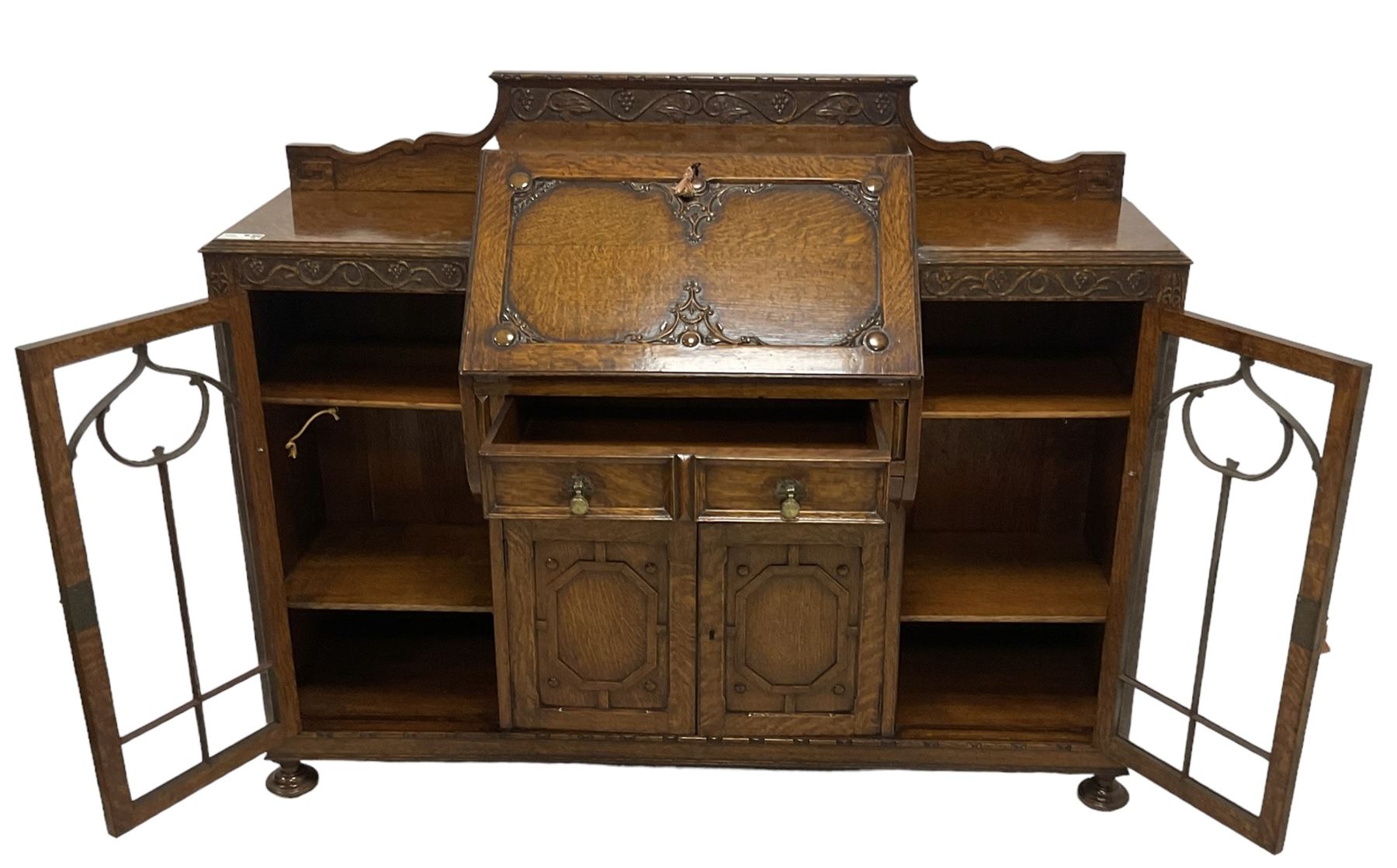 Early 20th century heavily carved oak bookcase bureau, raised back with grape-vine carved frieze, over fall-front enclosing fitted interior, above two short drawers and curved frieze drawer, flanked by astragal glazed and panelled cupboards, on turned feet