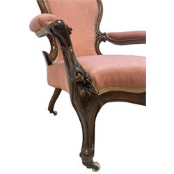 Victorian mahogany framed open armchair, spoon-back and sprung seat upholstered in pink fabric, scrolled arm terminals over serpentine fronted seat decorated with moulded carved floral motifs, cabriole supports with ceramic castors