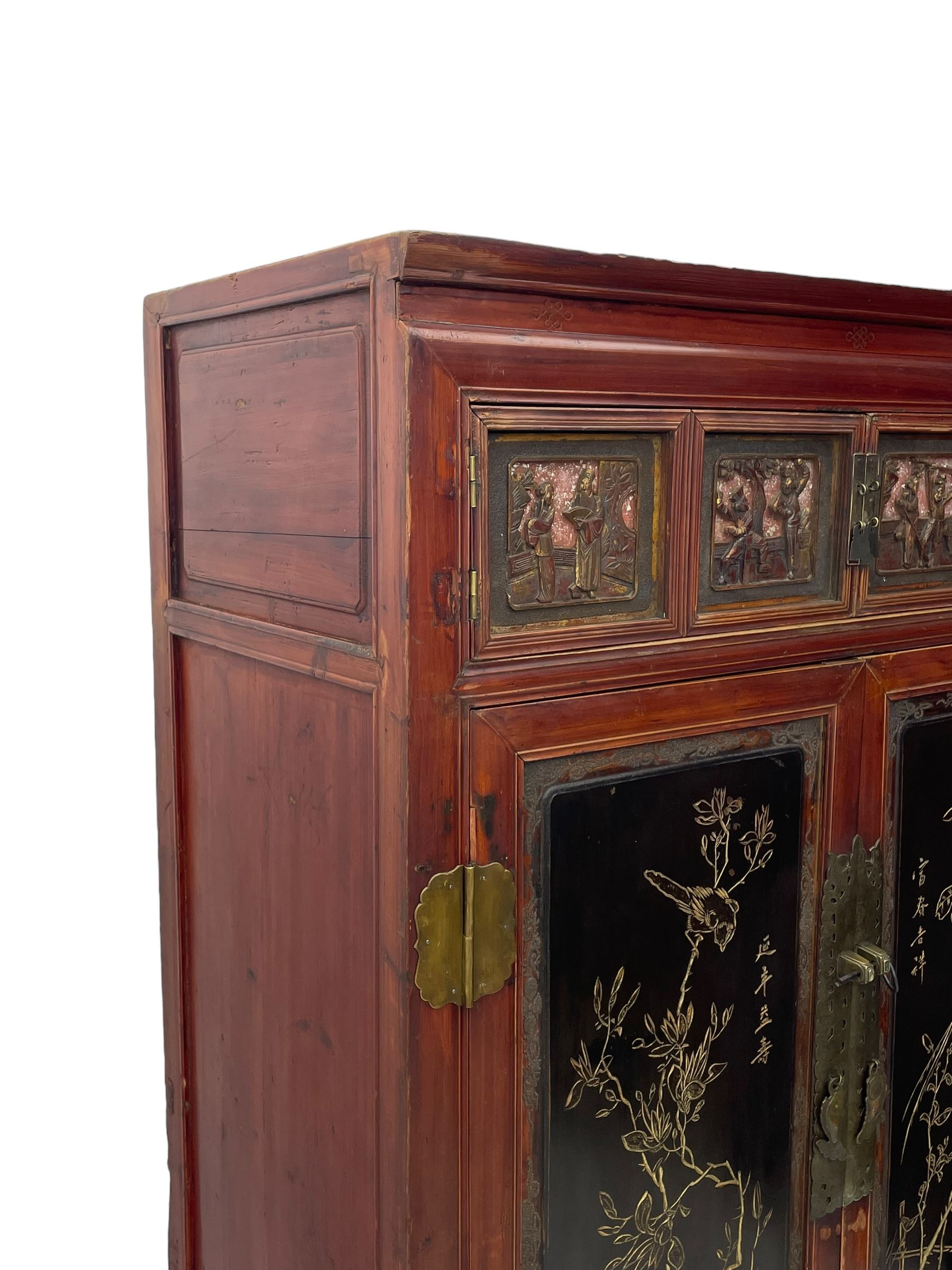 Late 19th century Chinese Qing dynasty red and black lacquered wood cabinet, Fujianese province, upper cupboard enclosed by two doors with relief carved and gilt panels depicting figures behind glass, the large cupboard enclosed by two panelled doors carved with trailing foliate branches and birds, inscribed with Chinese characters, to the left ‘Prolong life’ and to the right ‘Wealth and good fortune’, fitted with two drawers, square supports with carved brackets 