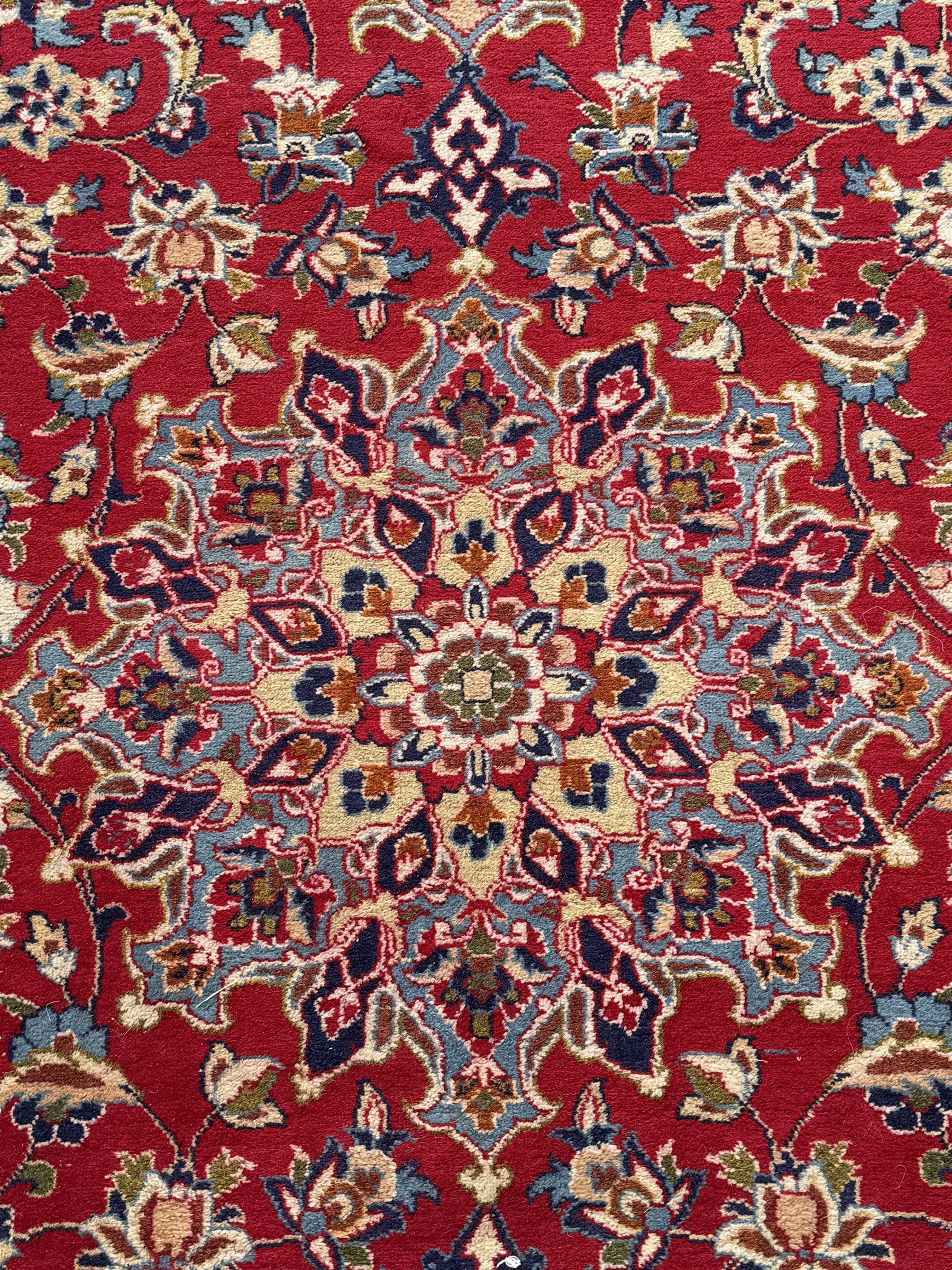 Persian Kashan red ground rug, the field decorated with a central floral medallion surrounded buy scrolling vine motifs and palmettes, the main border featuring a series of stylised floral motifs against a dark blue ground, enclosed by multiple guard stripes with alternating floral and geometric designs