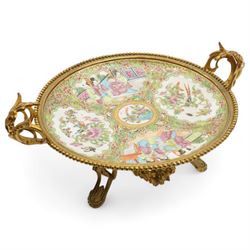 19th century Chinese Canton famille rose circular dish, painted with figures and exotic birds in shaped panels, against a floral and scroll ground, mounted in ormolu style gilt metal twin handled mount with beaded border and three scroll feet, W30cm x H14cm