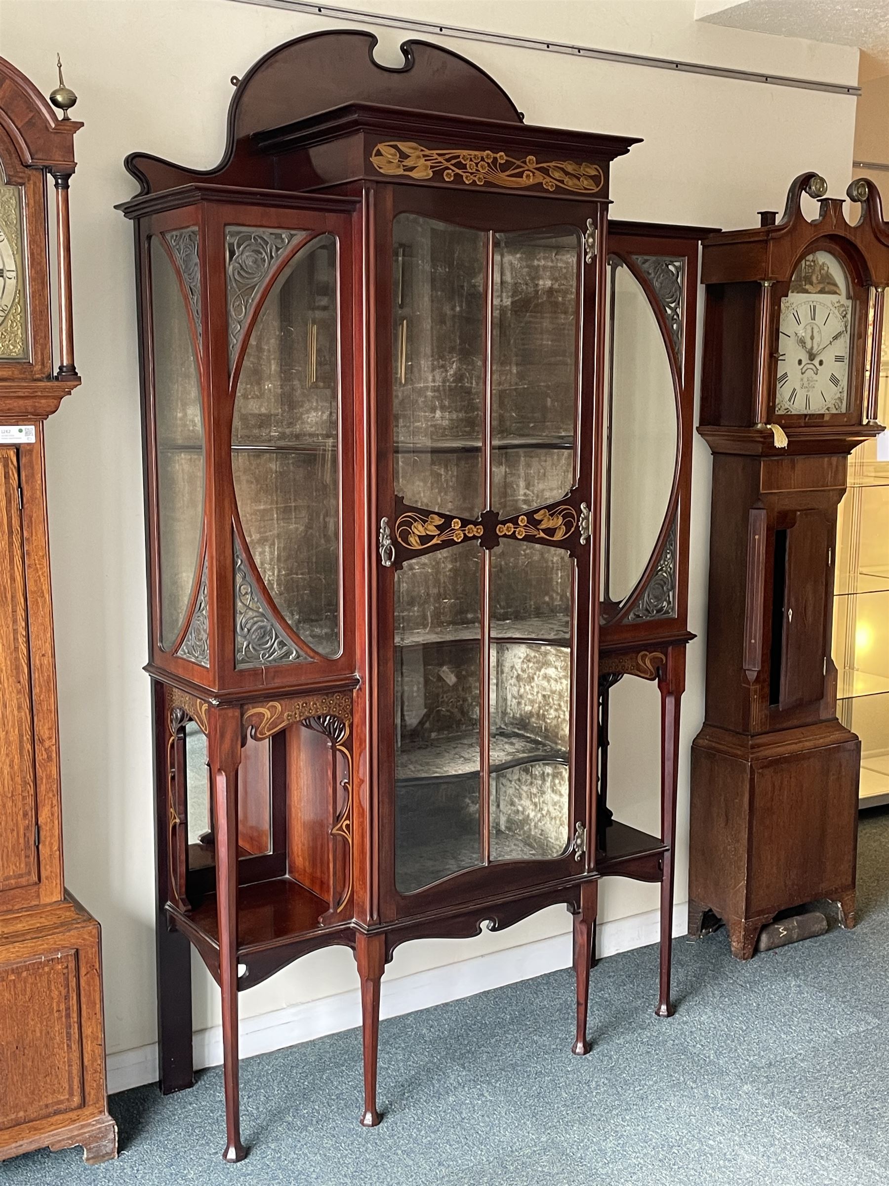 Art Nouveau period inlaid mahogany display cabinet, raised shaped and moulded pediment over projecting moulded cornices, the upper frieze inlaid with trailing branches and tulips, single astragal and bevel glazed door enclosed velvet lined interior fitted with shelves, flanked by curved glass panes decorated with pierced metal foliate spandrels, lower flanking shelves with mirror backs enclosed within shaped and pierced rails inlaid with flower heads, on tapering octagonal supports terminating to compressed pad feet, the lock stamped 'Salmon Bros, London' 