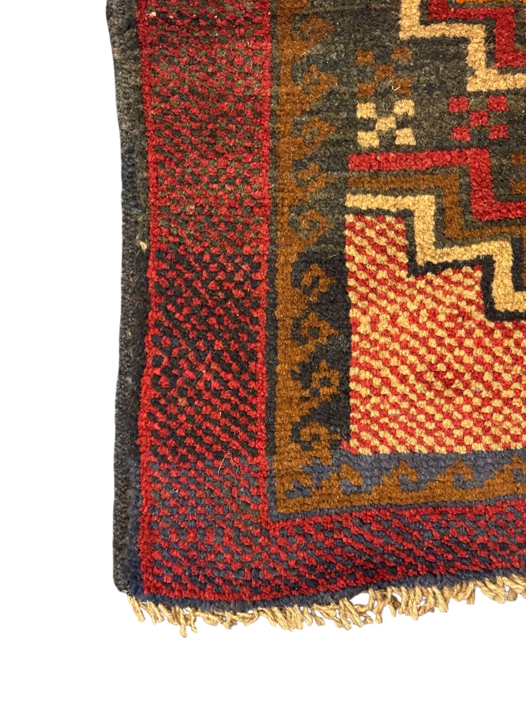 Baluchi crimson ground rug, the central field decorated with two stacked geometric lozenge medallions, surrounded by stepped multicoloured motifs, the wide border with alternating angular motifs in deep red, within a narrow brown guard stripe