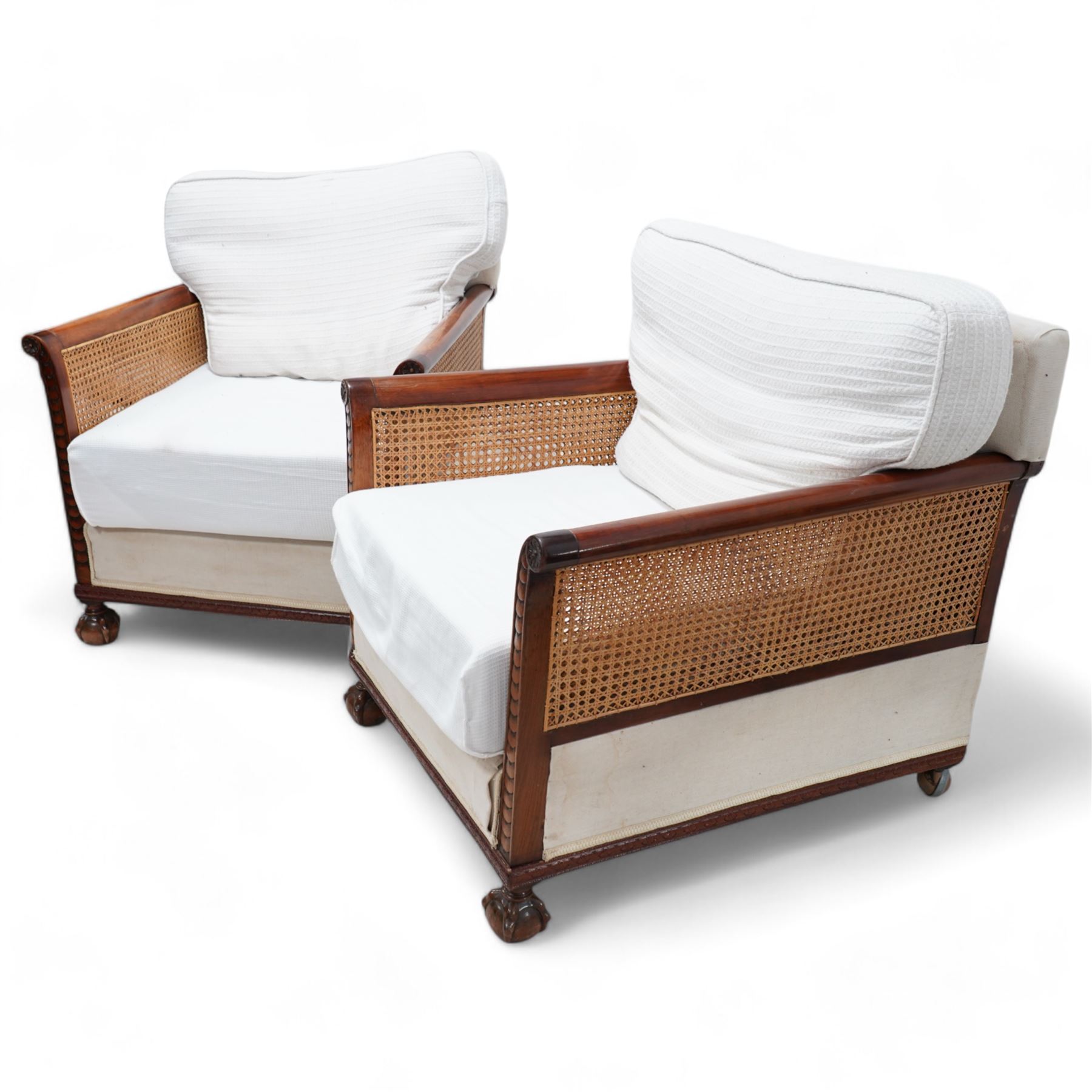 Pair of early 20th century mahogany framed bergère armchairs, upholstered back and seat flanked by rolled double-caned arms with carved scale decoration, floral and ribbon-twist carved lower edge, over ball-and-claw feet