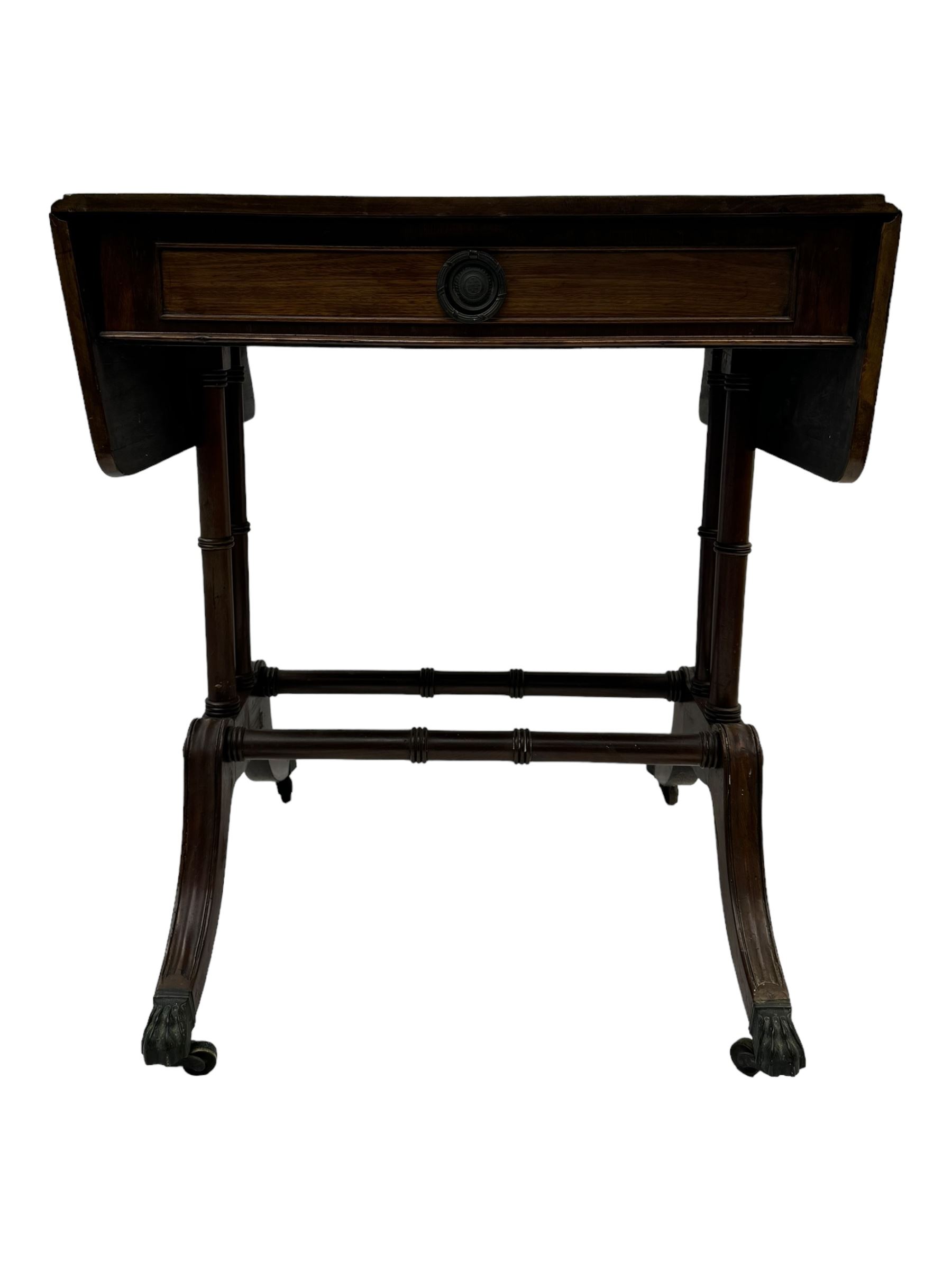 Mahogany drop-leaf sofa table, rectangular top over single frieze drawer with carved rosette handle, twin turned columns united by twin stretchers, on splayed supports terminating in paw feet with castors