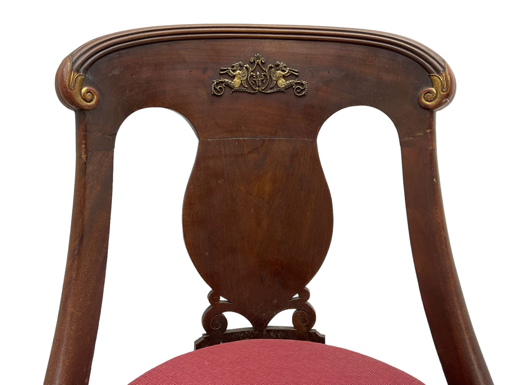 Pair of French Empire mahogany spoon0back side chairs, reeded cresting rail with scrolled ends over a shaped and pierced back splat, decorated with gilt metal mount in the form of winged classical maidens playing panpipes, drop-in seat upholstered in crimson lozenge patterned fabric flanked by gilt stylised scaled fish mounts, the apron with neoclassical gilt metal mounts, raised on sabre supports with paw feet