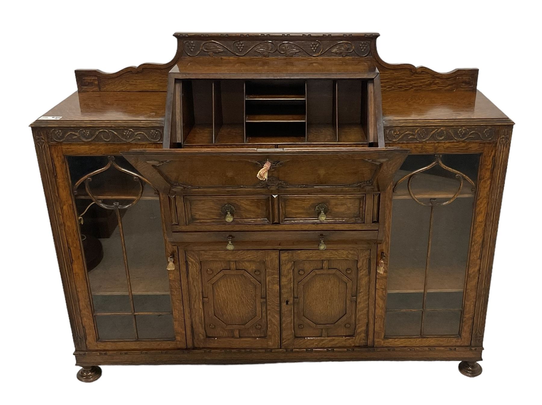 Early 20th century heavily carved oak bookcase bureau, raised back with grape-vine carved frieze, over fall-front enclosing fitted interior, above two short drawers and curved frieze drawer, flanked by astragal glazed and panelled cupboards, on turned feet