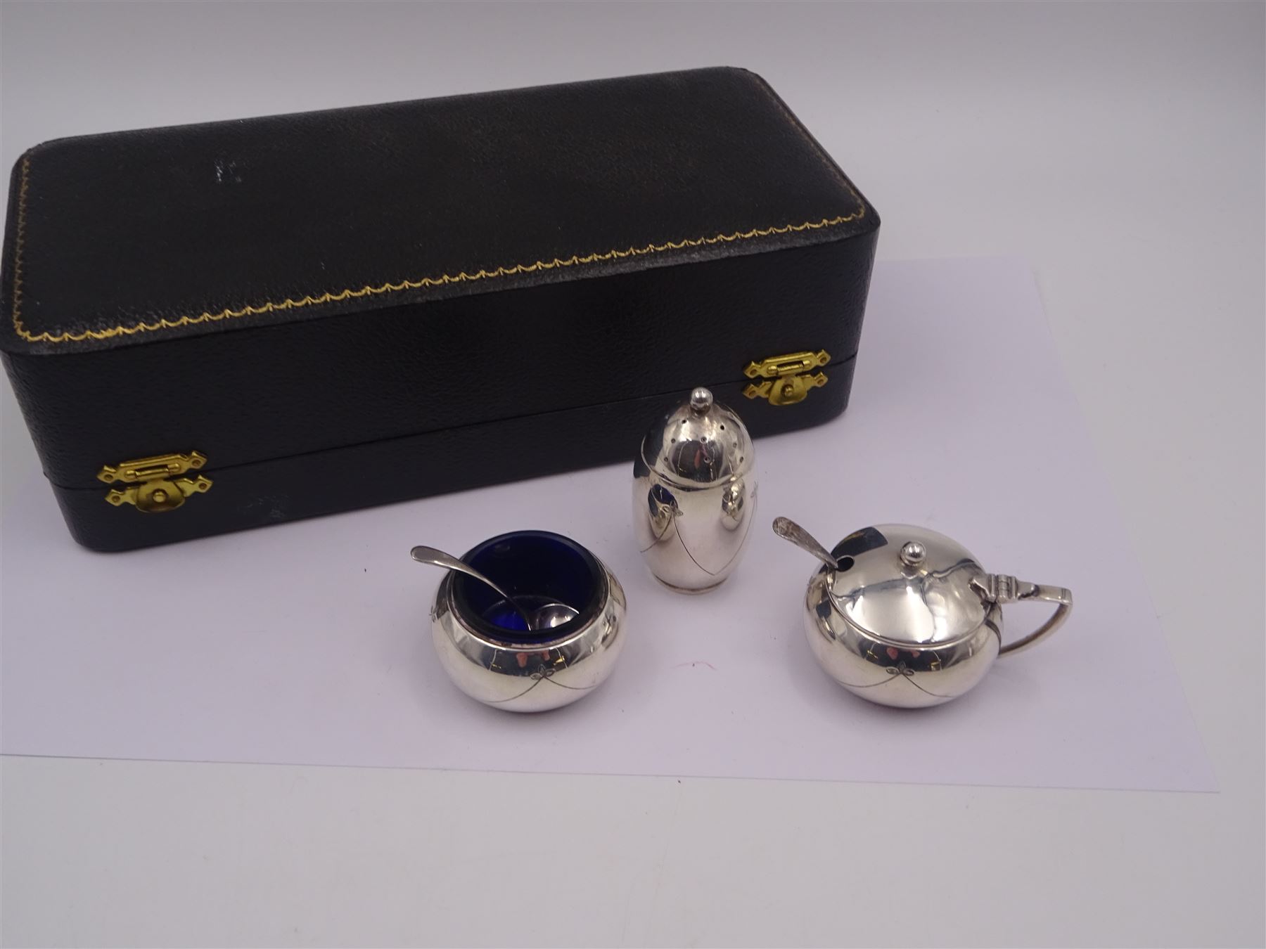 1950s silver three piece cruet set, comprising pepper shaker, open salt, mustard pot and cover, engraved with ribbon swags, hallmarked Joseph Gloster Ltd, Birmingham 1956, salt and mustard pots with blue glass liners, together with two condiment spoons, hallmarked, all within a fitted silk and velvet lined case 